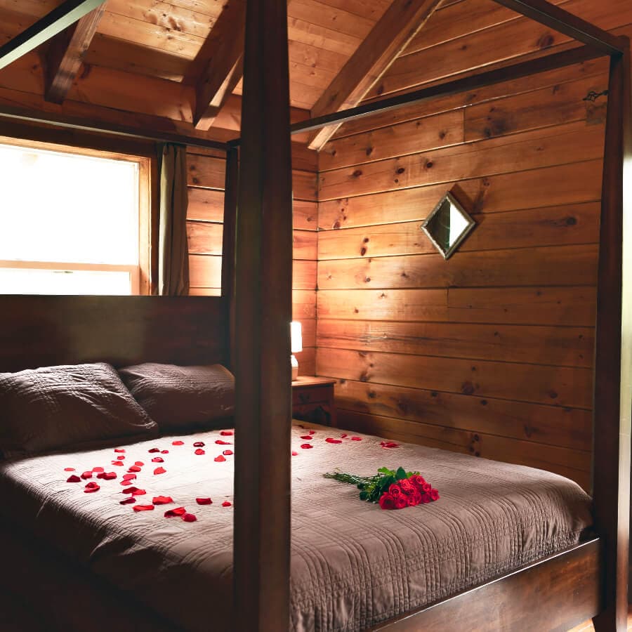 A cozy wooden bedroom with a four-poster bed adorned with rose petals and a bouquet of red roses.