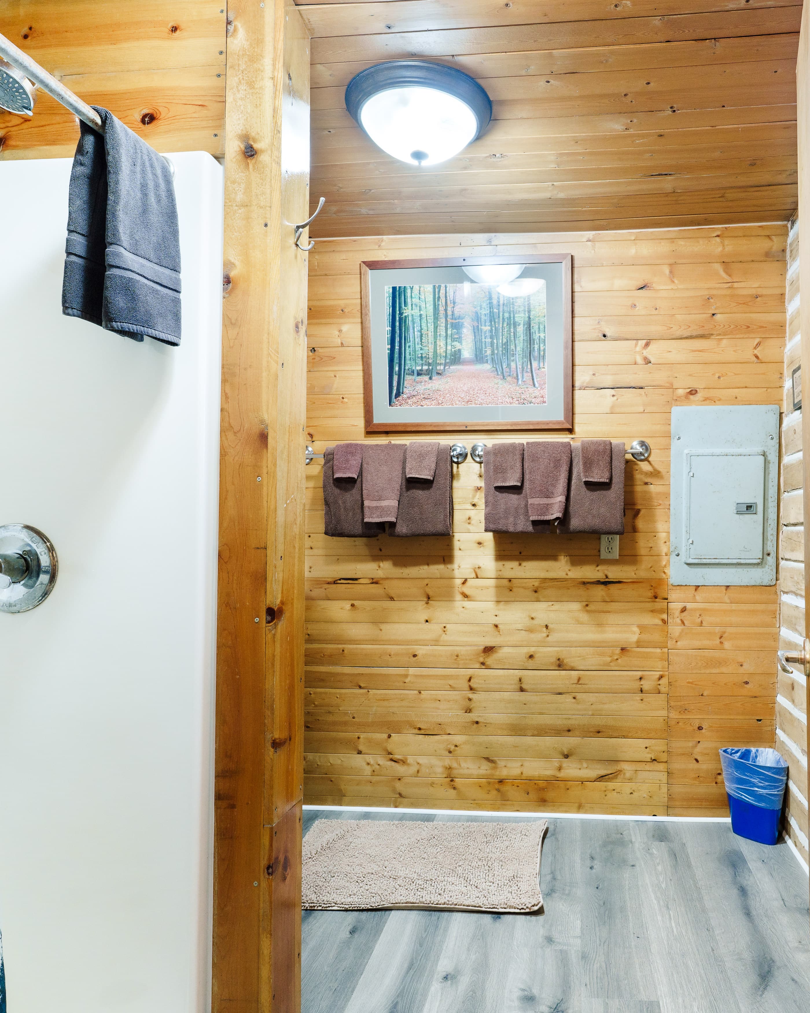 A wooden-paneled bathroom featuring a shower, hanging brown towels, a nature-themed picture, and a small blue trash can.
