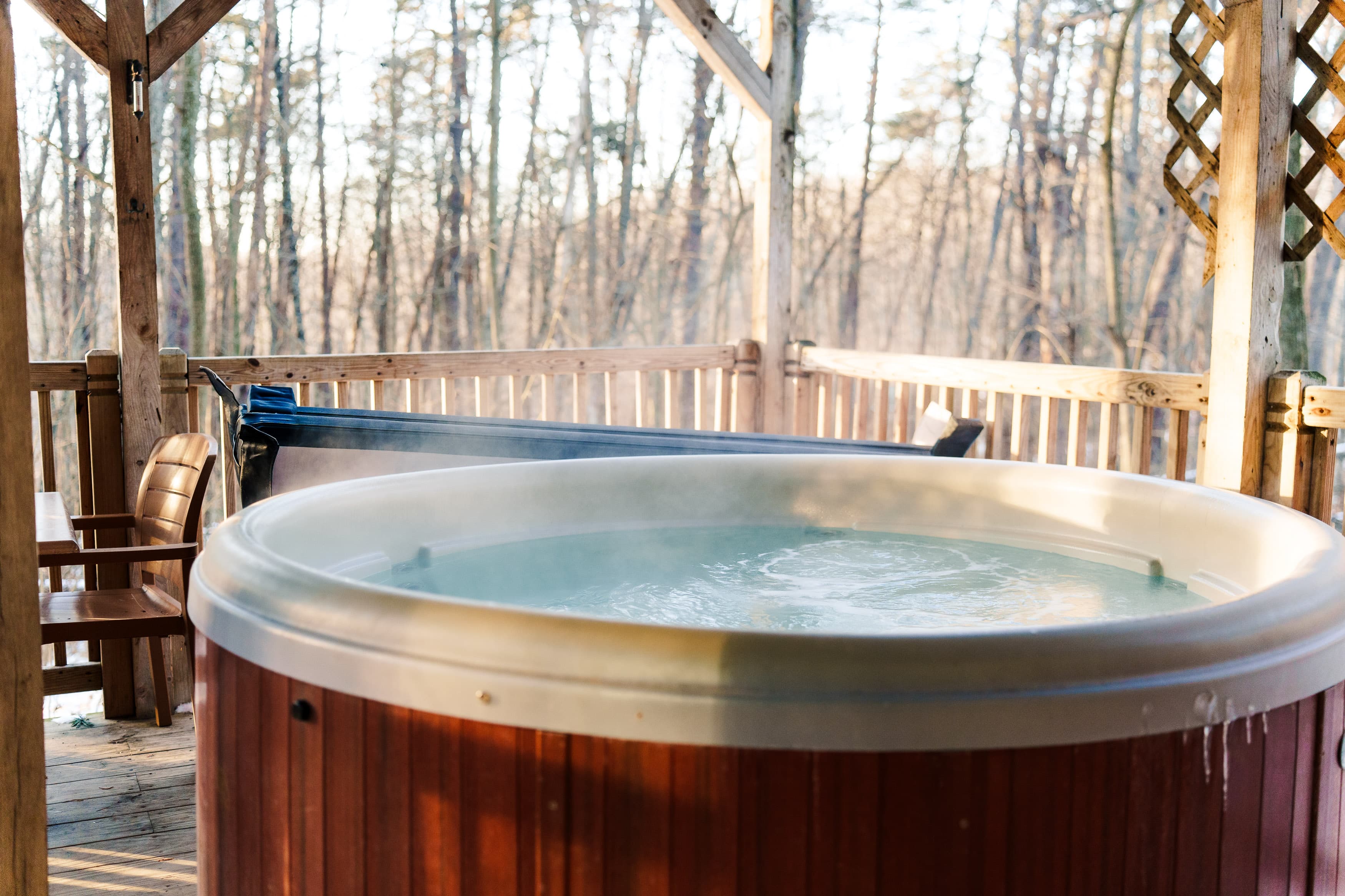 A wooden hot tub steams gently on a deck surrounded by trees.