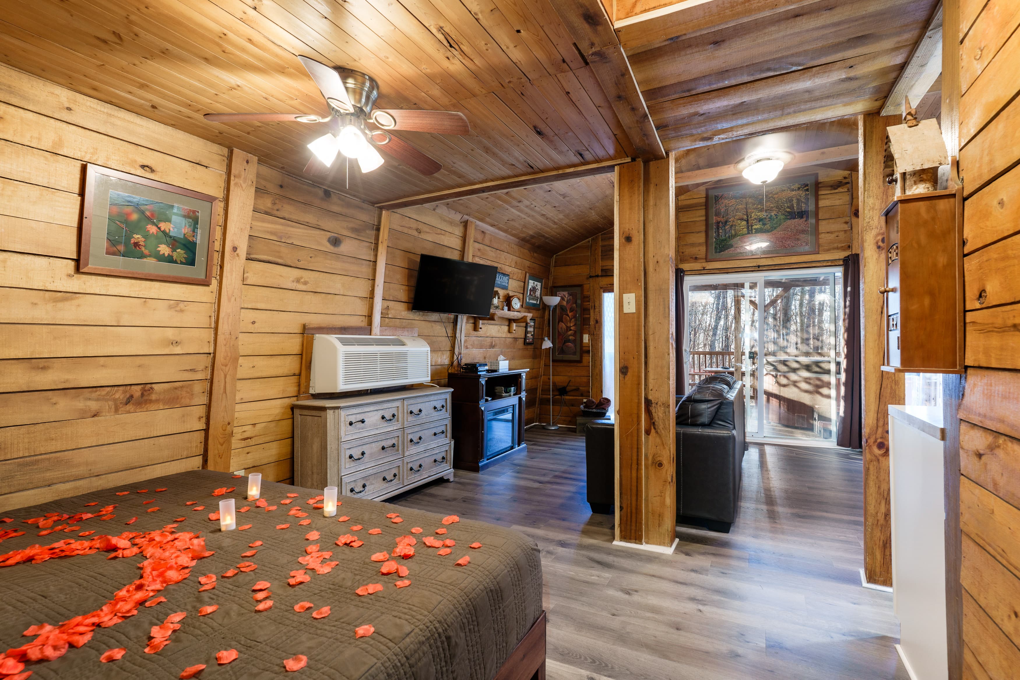 A rustic, cozy bedroom featuring wooden walls, a fan, roses on the bed, and a view of an outdoor deck.
