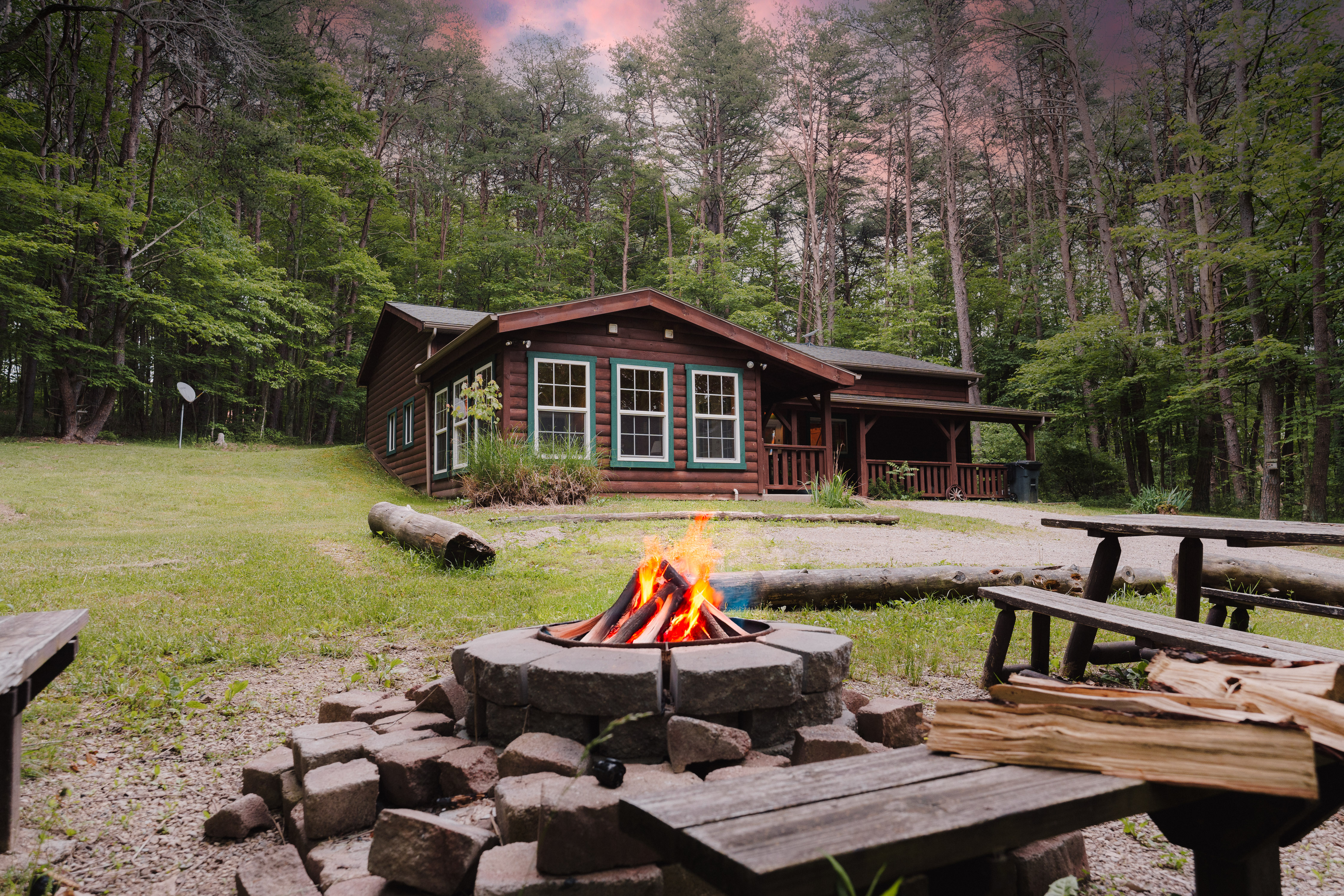 A rustic wooden cabin with teal-trimmed windows nestled in a forest, viewed past a burning stone fire pit, chopped wood, and picnic benches in the foreground.