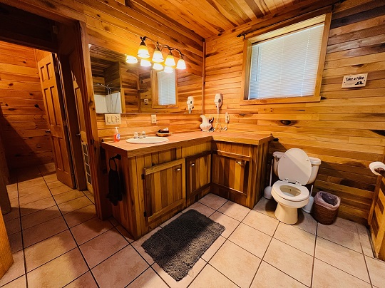 Rustic bathroom with wood-paneled walls and ceiling, featuring an L-shaped wooden vanity with a sink and vanity lights, a large mirror, tile flooring, and a white toilet next to a window with blinds.