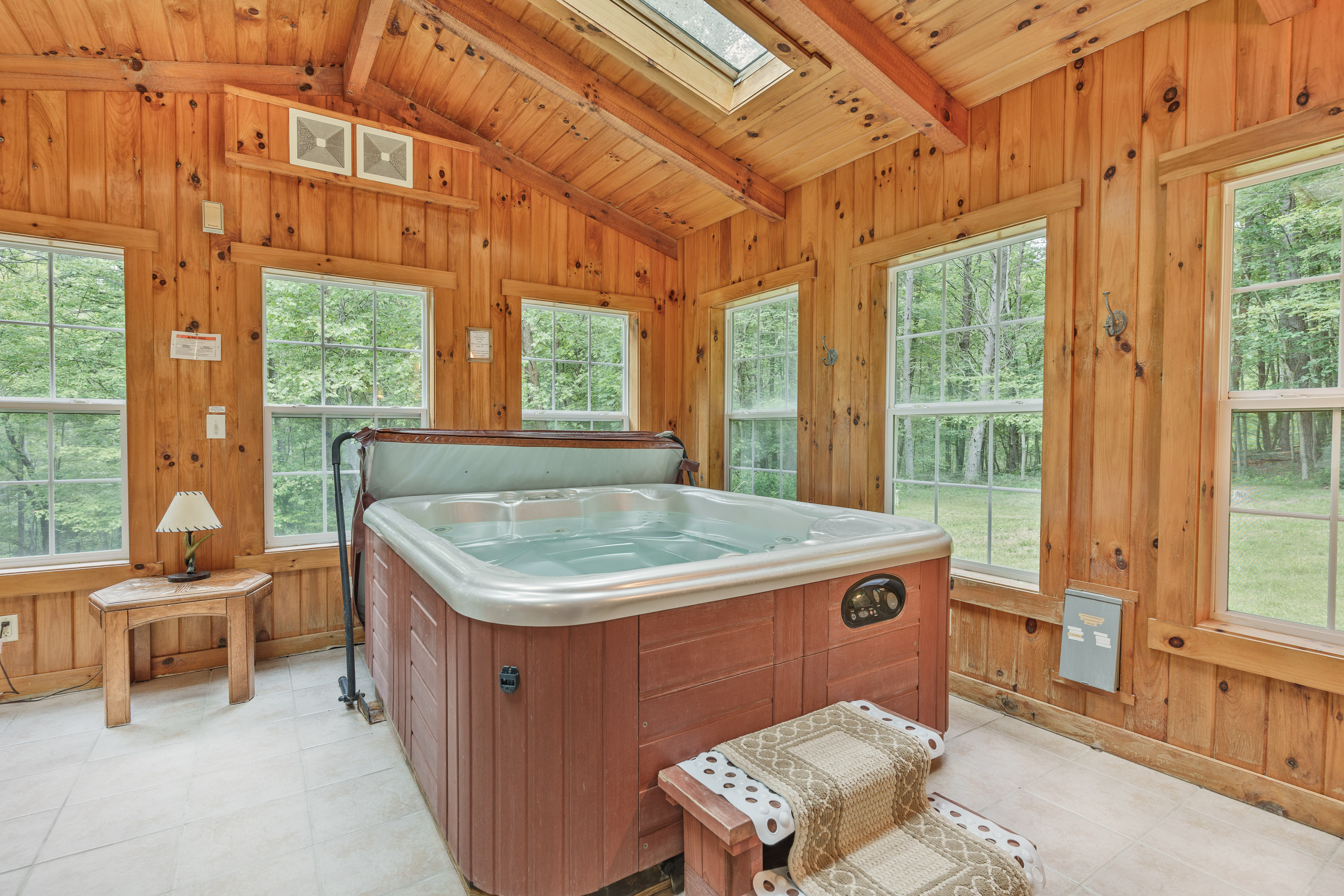 Square indoor hot tub located in a bright, wood-paneled sunroom featuring a vaulted ceiling with skylights and wrap-around windows overlooking the forest.