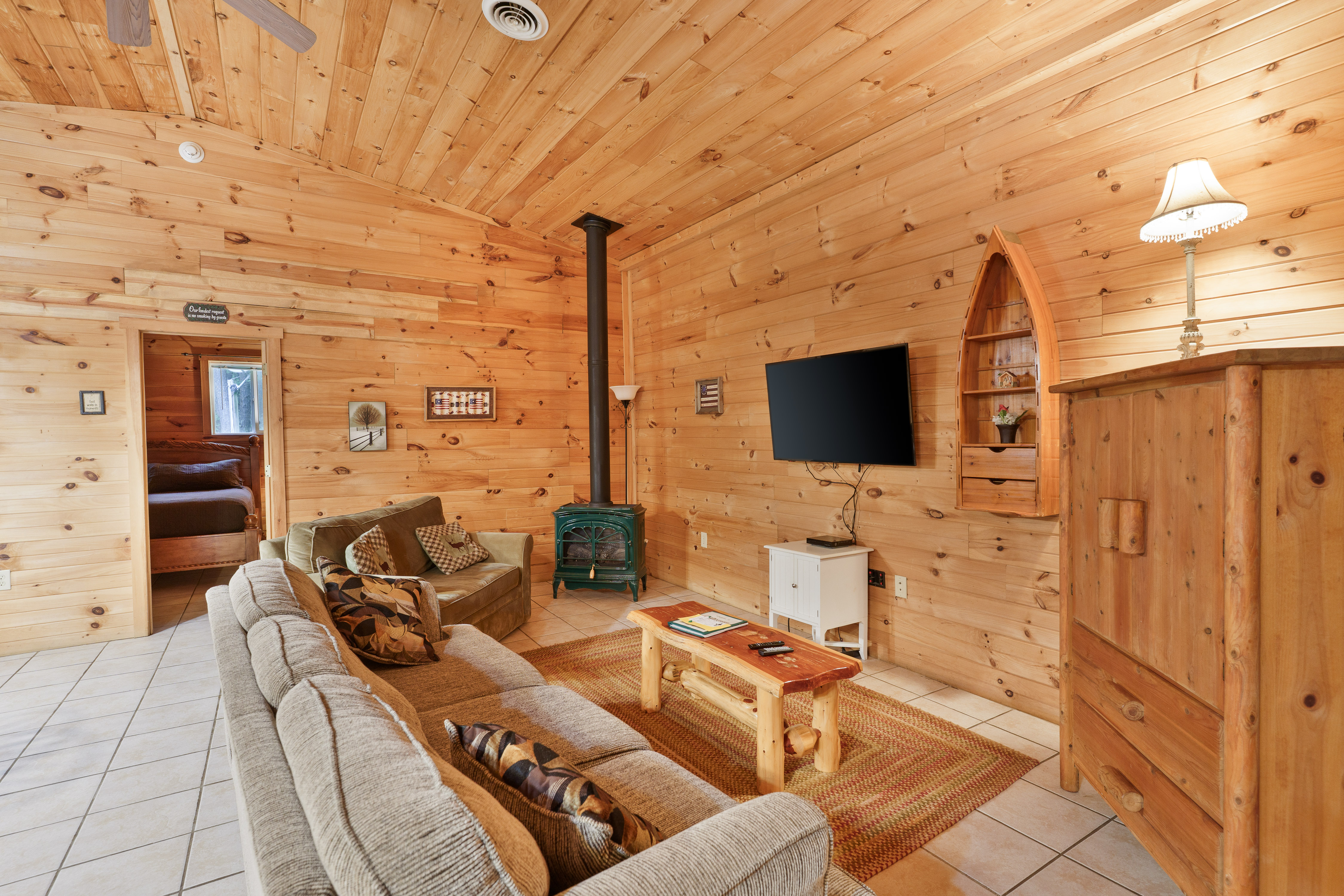 Spacious cabin living room with light wood paneling and vaulted ceilings, featuring two beige sofas, a rustic coffee table, a green wood-burning stove, a wall-mounted TV, and a decorative boat-shaped shelf.