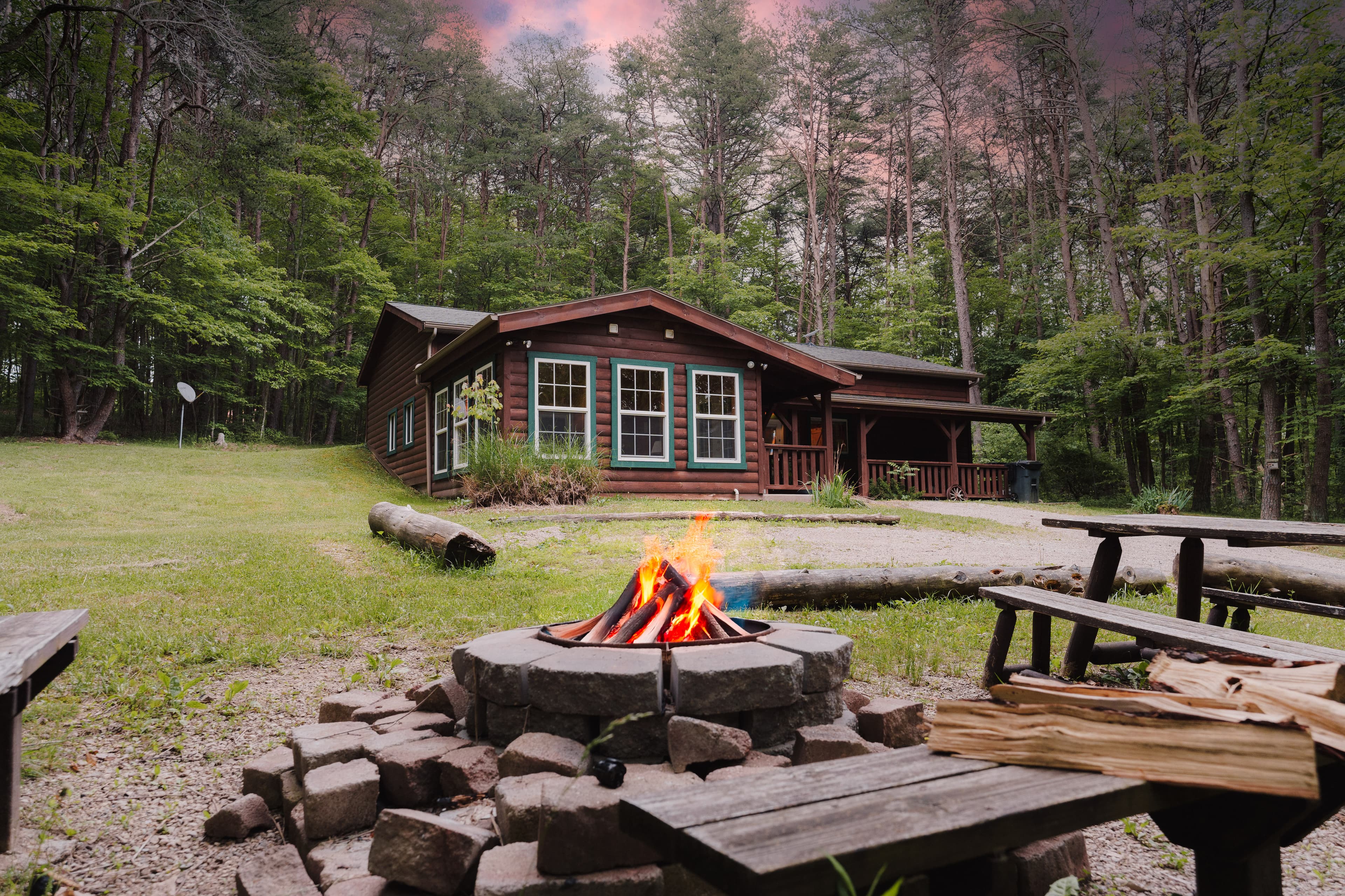 A rustic wooden cabin with teal-trimmed windows nestled in a forest, viewed past a burning stone fire pit, chopped wood, and picnic benches in the foreground.