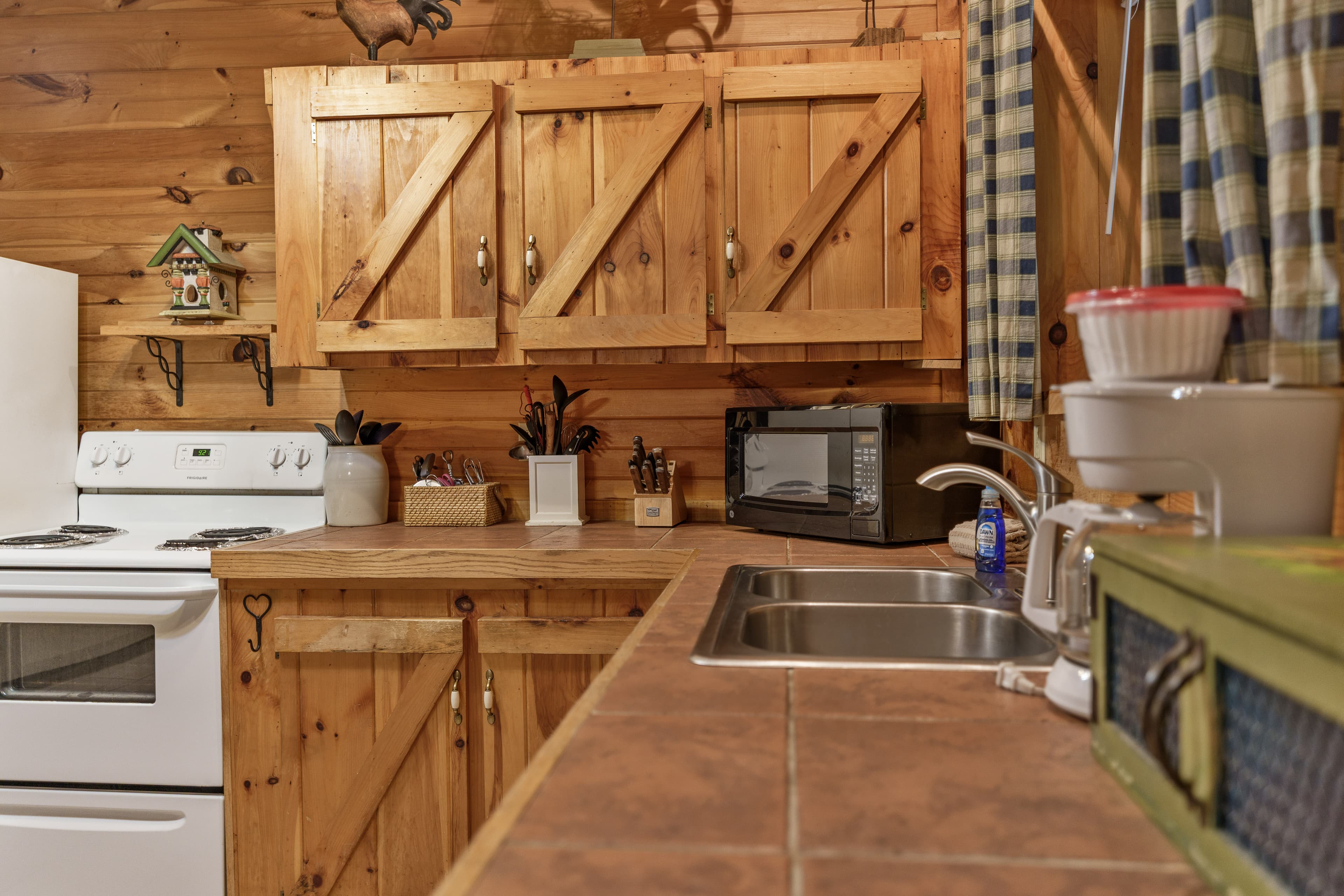 Close-up of a rustic kitchen corner featuring wooden cabinets with barn-style doors, brown tile countertops, and a double stainless steel sink, equipped with a white stove, microwave, coffee maker, and blue plaid curtains.