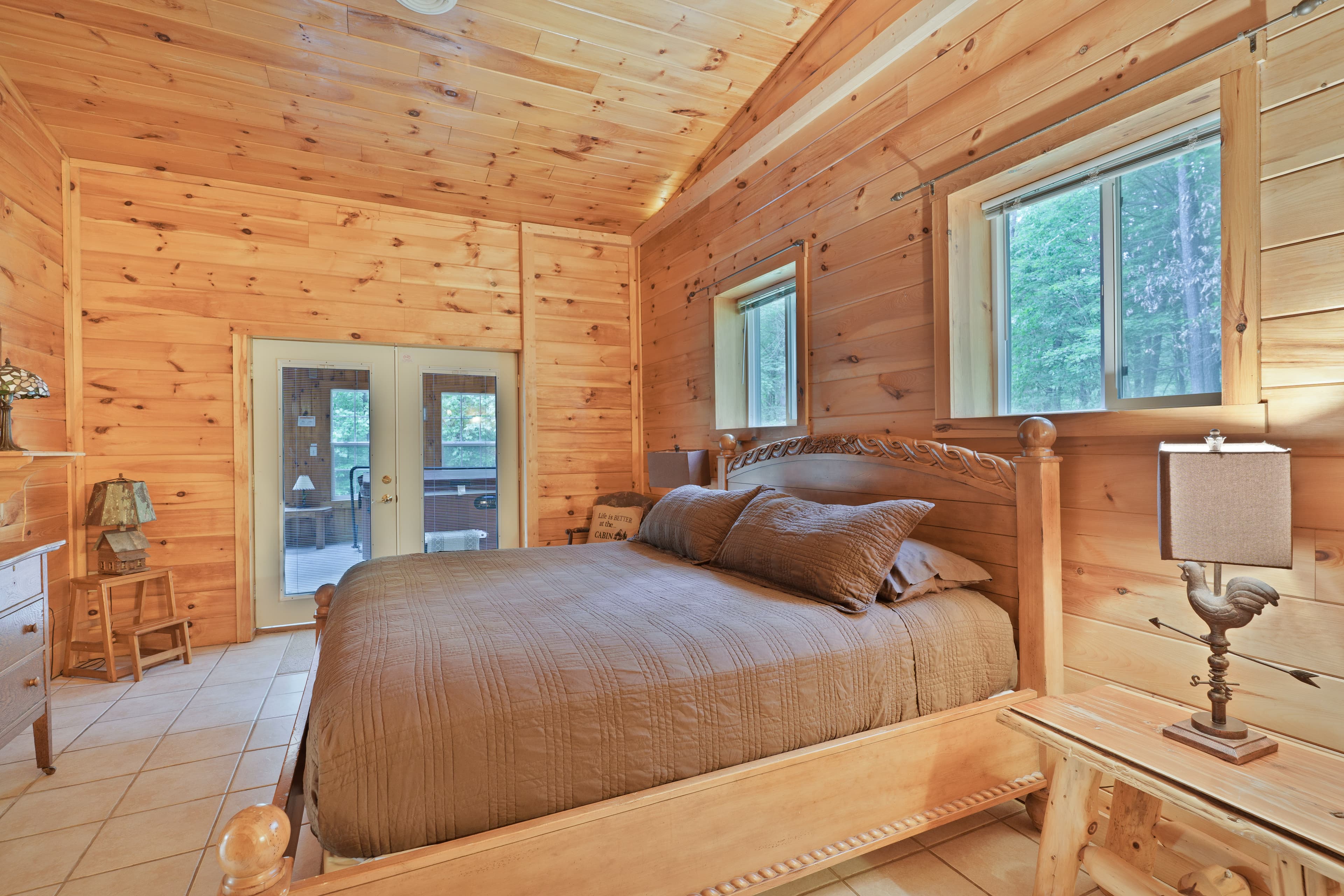 Bedroom inside a cabin with light pine wood paneling on the walls and vaulted ceiling, featuring a wooden bed with brown bedding, tile floors, and double glass doors leading to a porch.