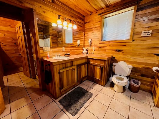 Rustic bathroom with wood-paneled walls and ceiling, featuring an L-shaped wooden vanity with a sink and vanity lights, a large mirror, tile flooring, and a white toilet next to a window with blinds.