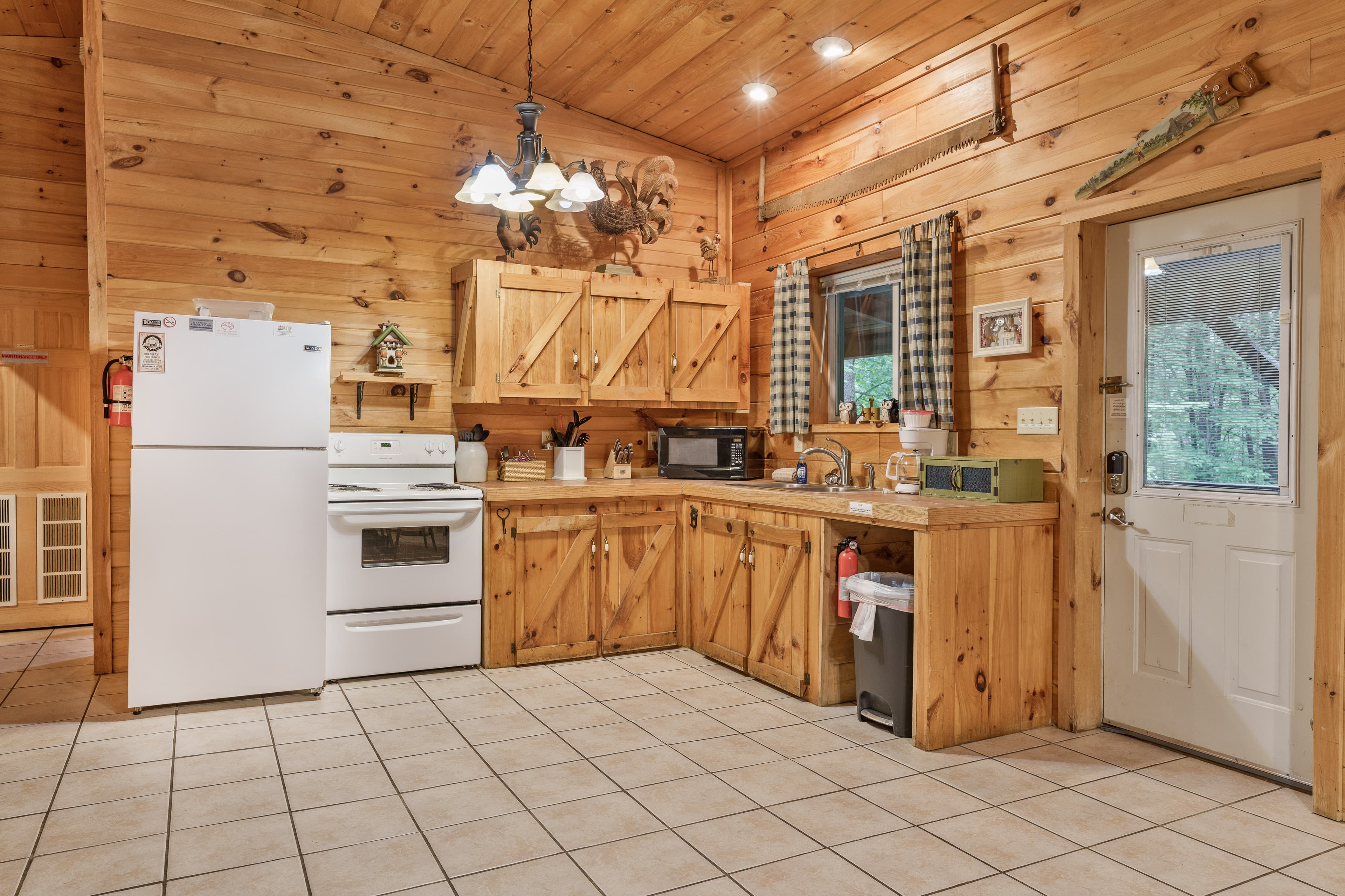 Rustic kitchen with light wood paneling on the walls and vaulted ceiling, featuring wooden cabinets with barn-style doors, white appliances including a refrigerator and stove, tile flooring, and a door leading outside.
