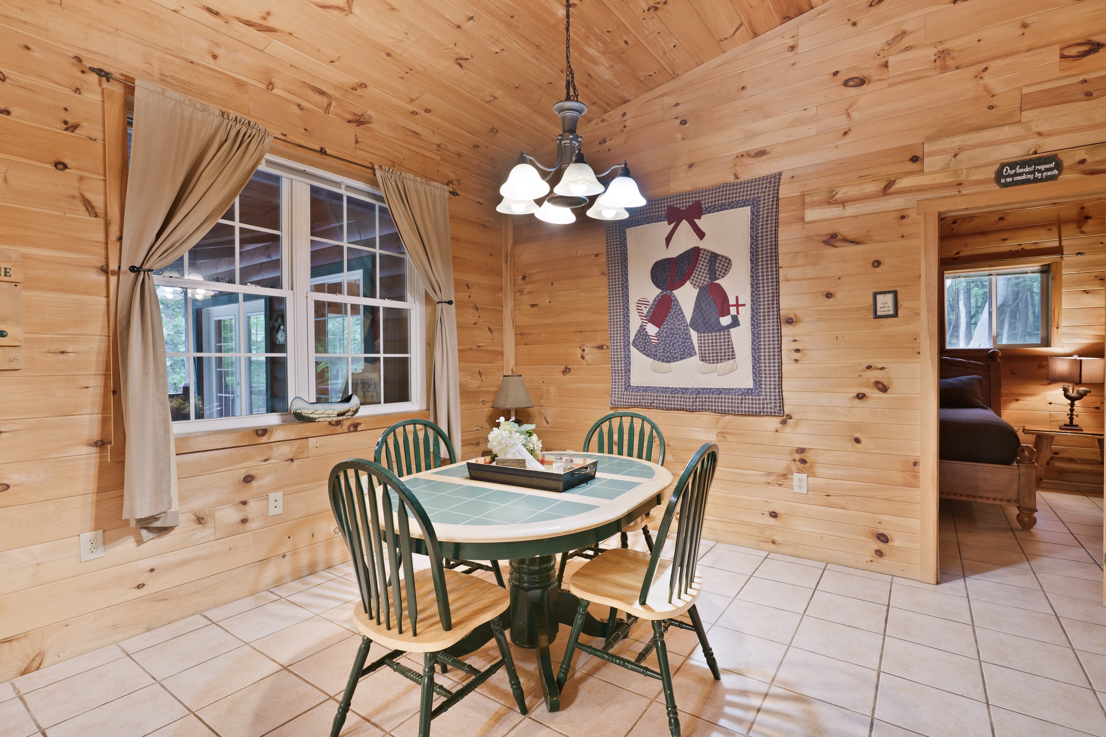 Dining area with light wood paneling and tile floors, featuring a round table with green chairs, a five-light chandelier, a decorative wall quilt, and a window with beige curtains.