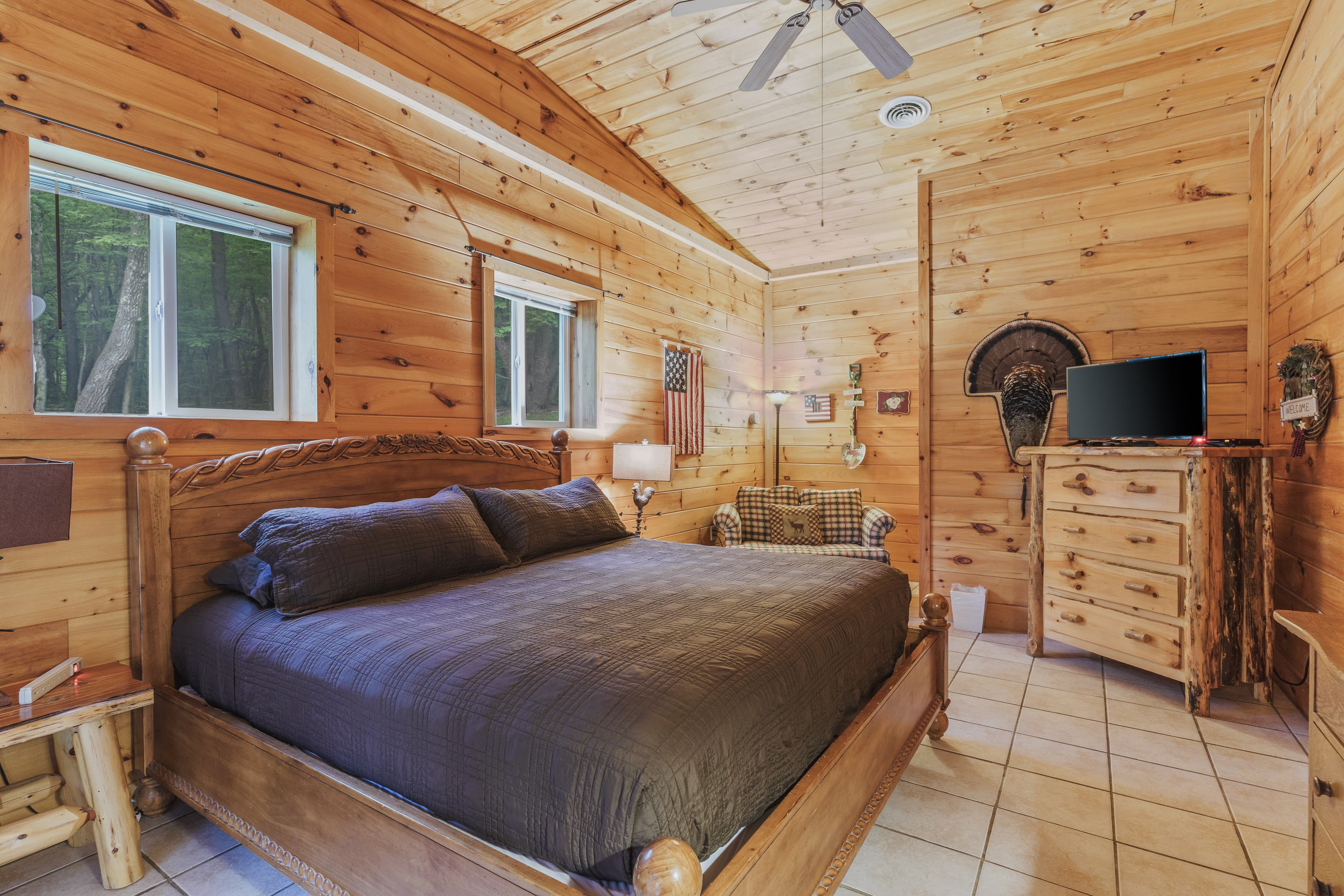 Rustic cabin bedroom with wood-paneled walls and a vaulted ceiling, featuring a large wooden bed with dark bedding, a log-style dresser with a TV, wild turkey wall decor, and tile flooring.