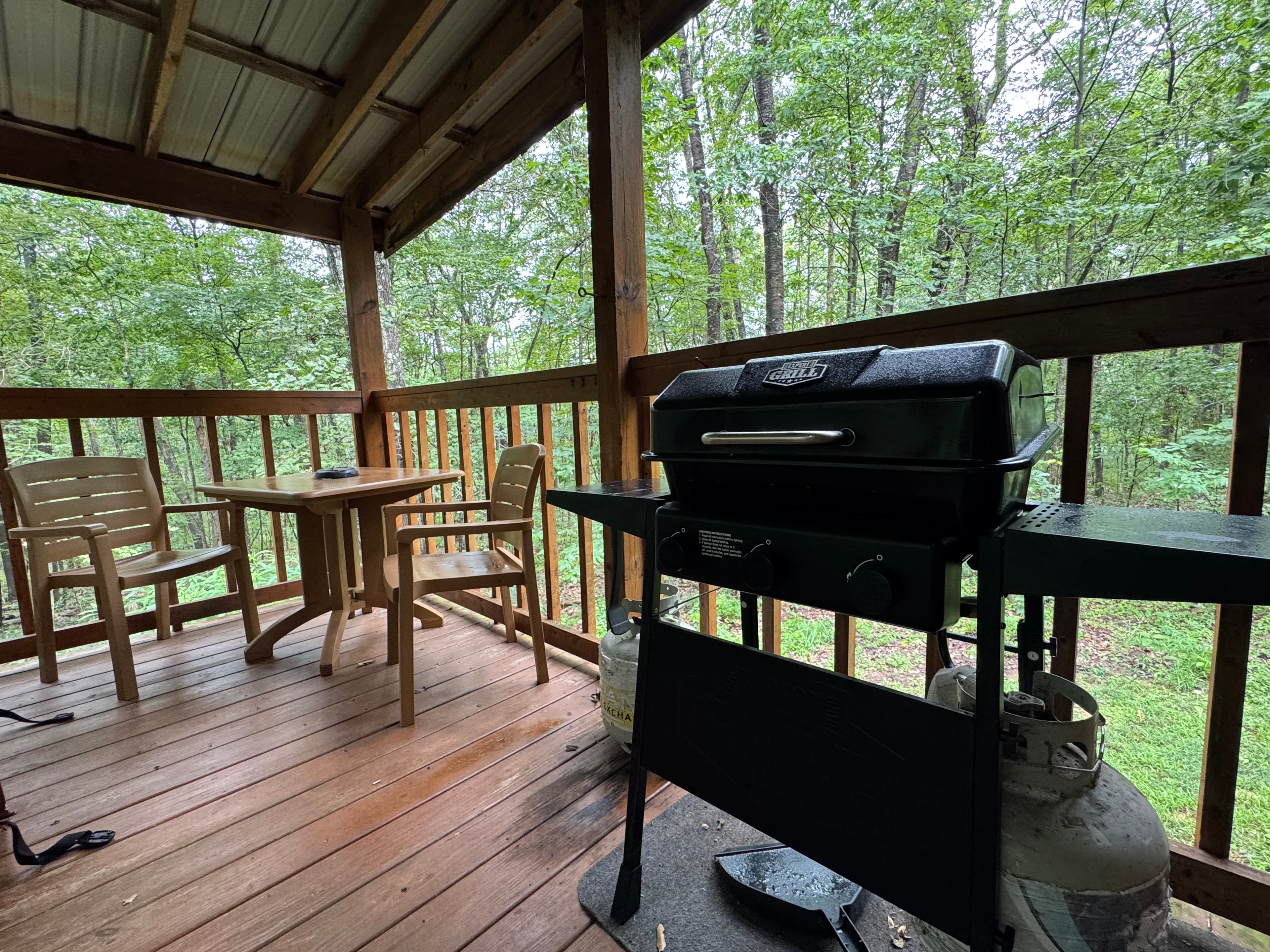 A gas grill sits on a wooden deck with two chairs and a table, surrounded by greenery.