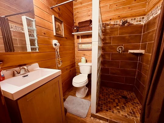 A cozy bathroom featuring wooden walls, a white sink, toilet, and a tiled shower.