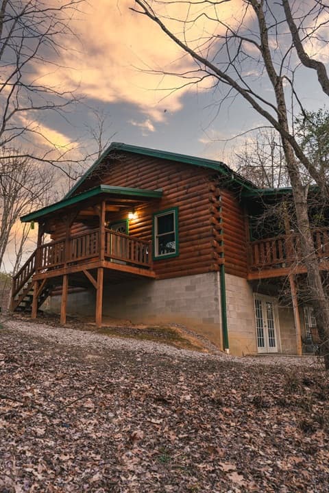 A cozy log cabin sits on a hillside surrounded by bare trees under a colorful sky.
