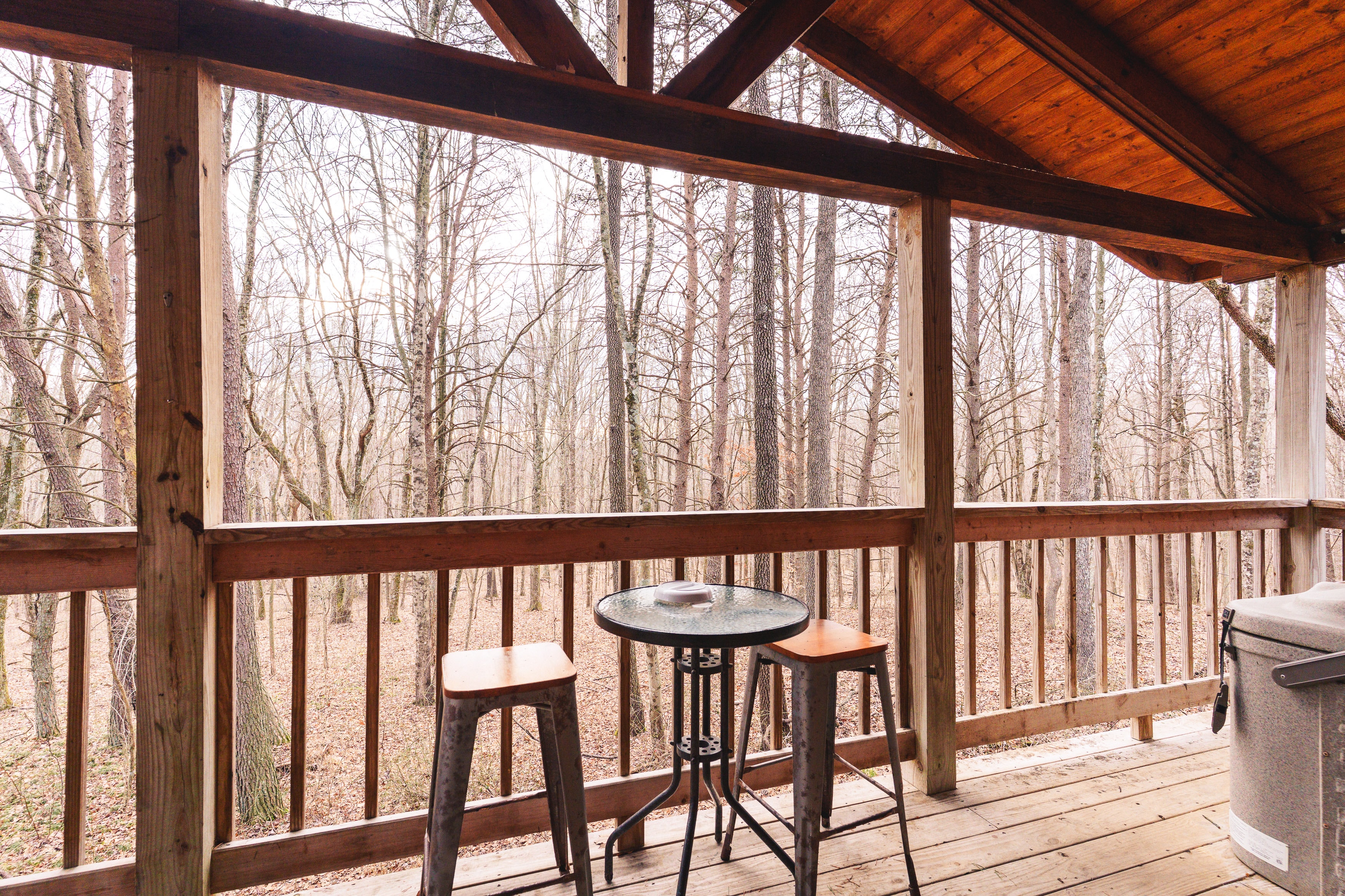 A cozy wooden balcony with a table and two chairs overlooks a serene, wooded area.