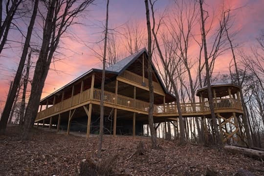 A modern wooden cabin nestled among bare trees under a colorful sunset sky.