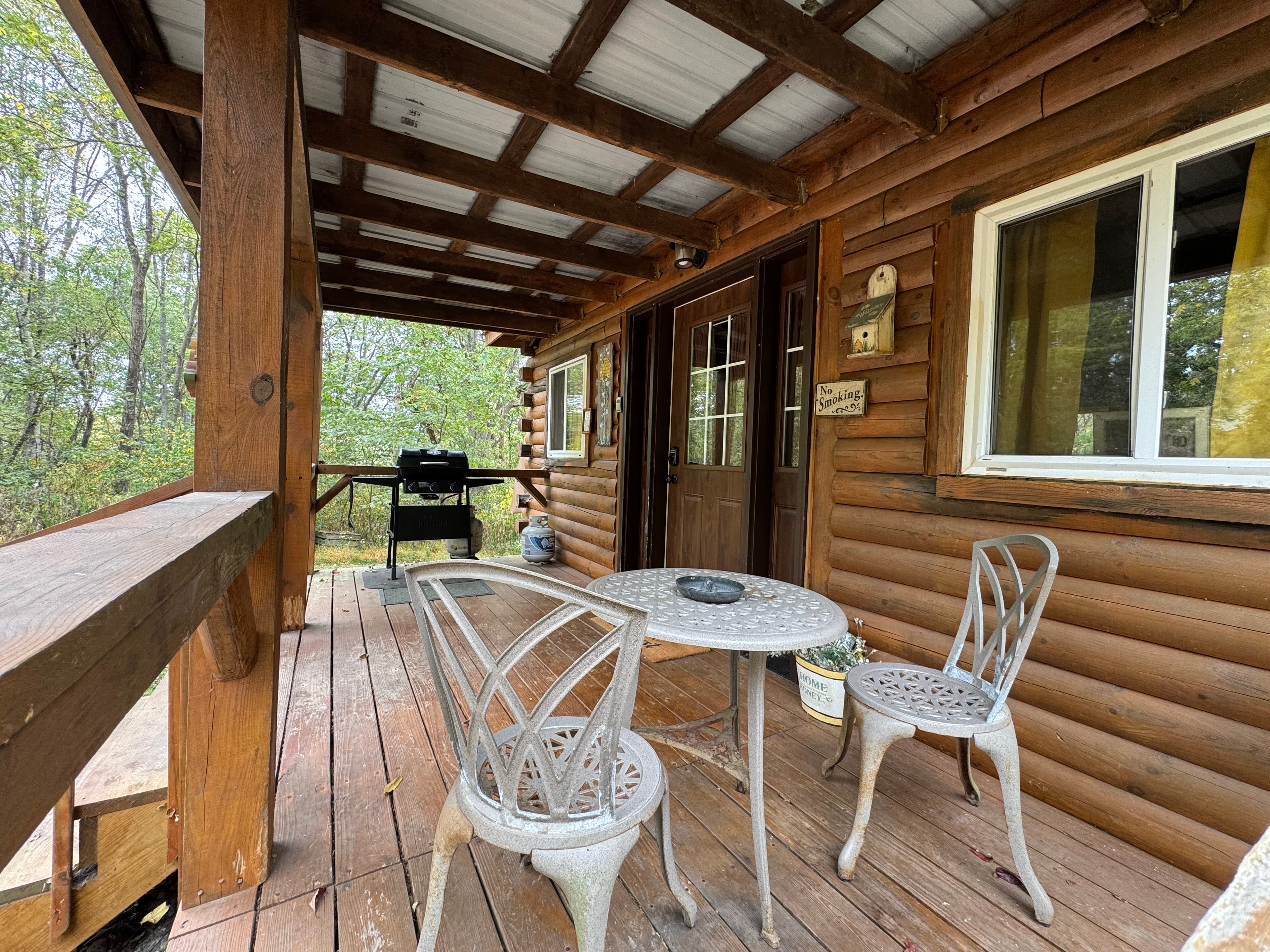 A rustic porch with a table and chairs, overlooking a wooded area.