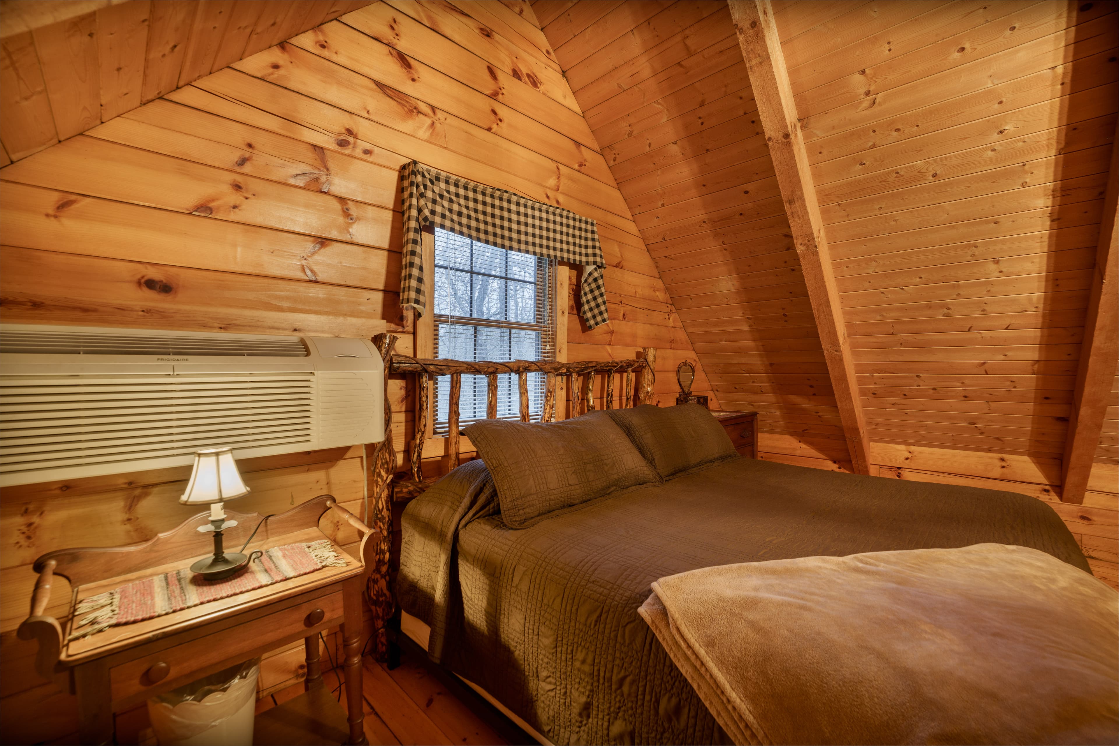 Cozy wooden bedroom with a bed, bedside table, lamp, and a window with plaid curtains.