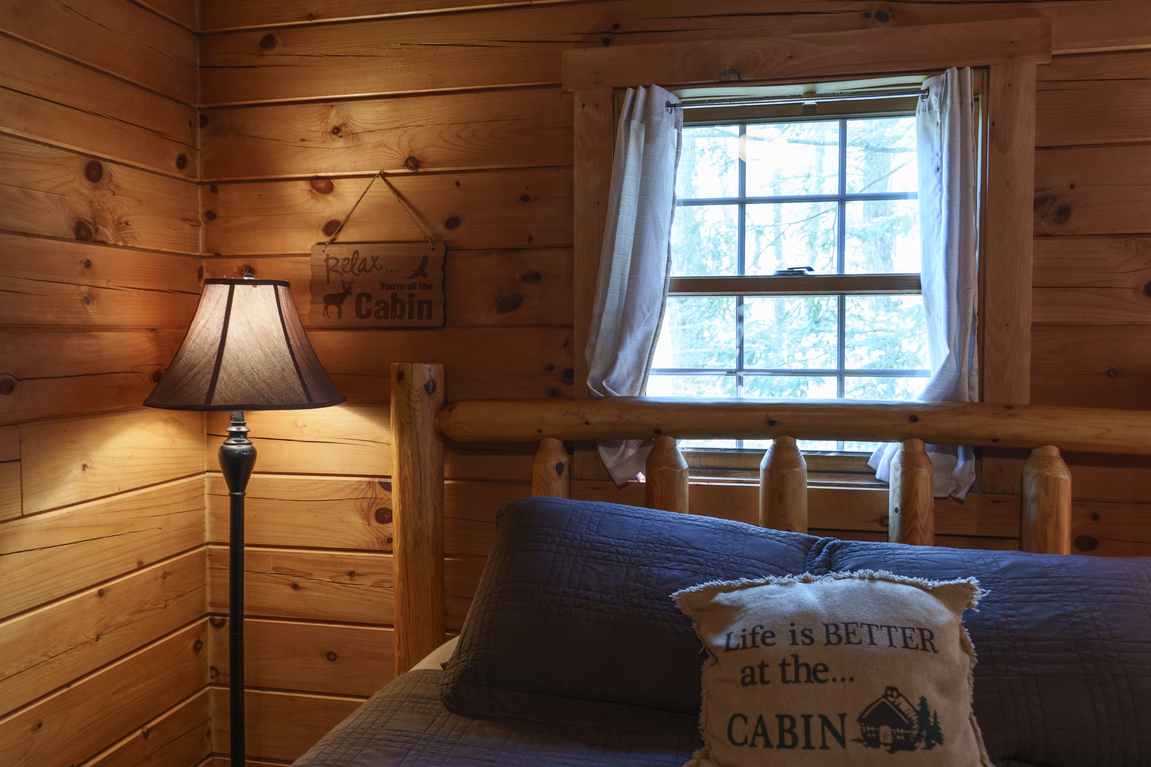 Cozy cabin bedroom with wooden walls, a lamp, and a window letting in natural light.