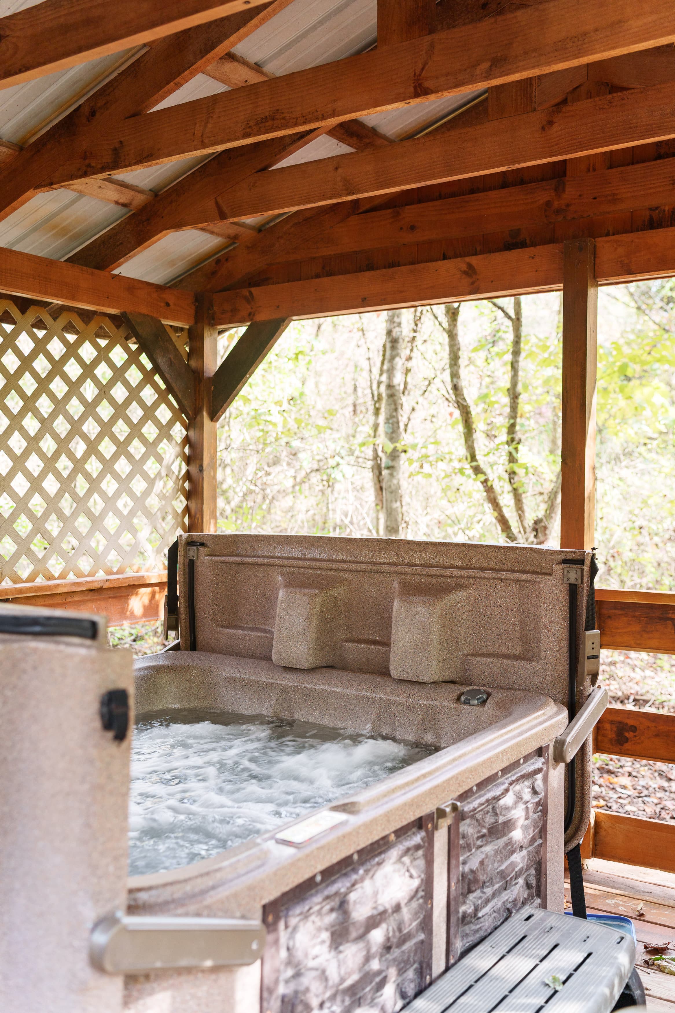 A hot tub with bubbling water situated under a wooden gazebo surrounded by trees.