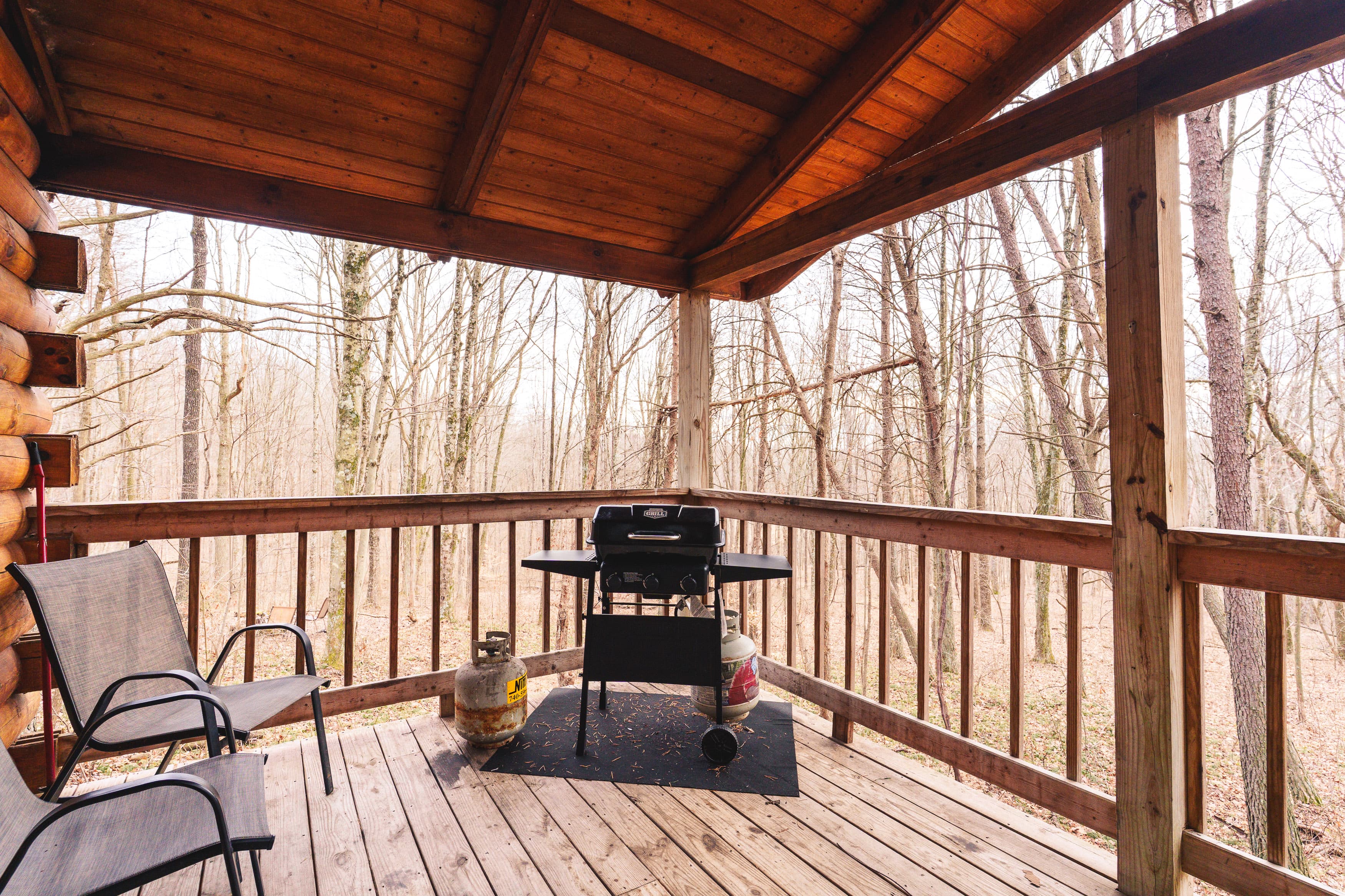 A covered wooden patio features a grill and two chairs, overlooking a wooded area.