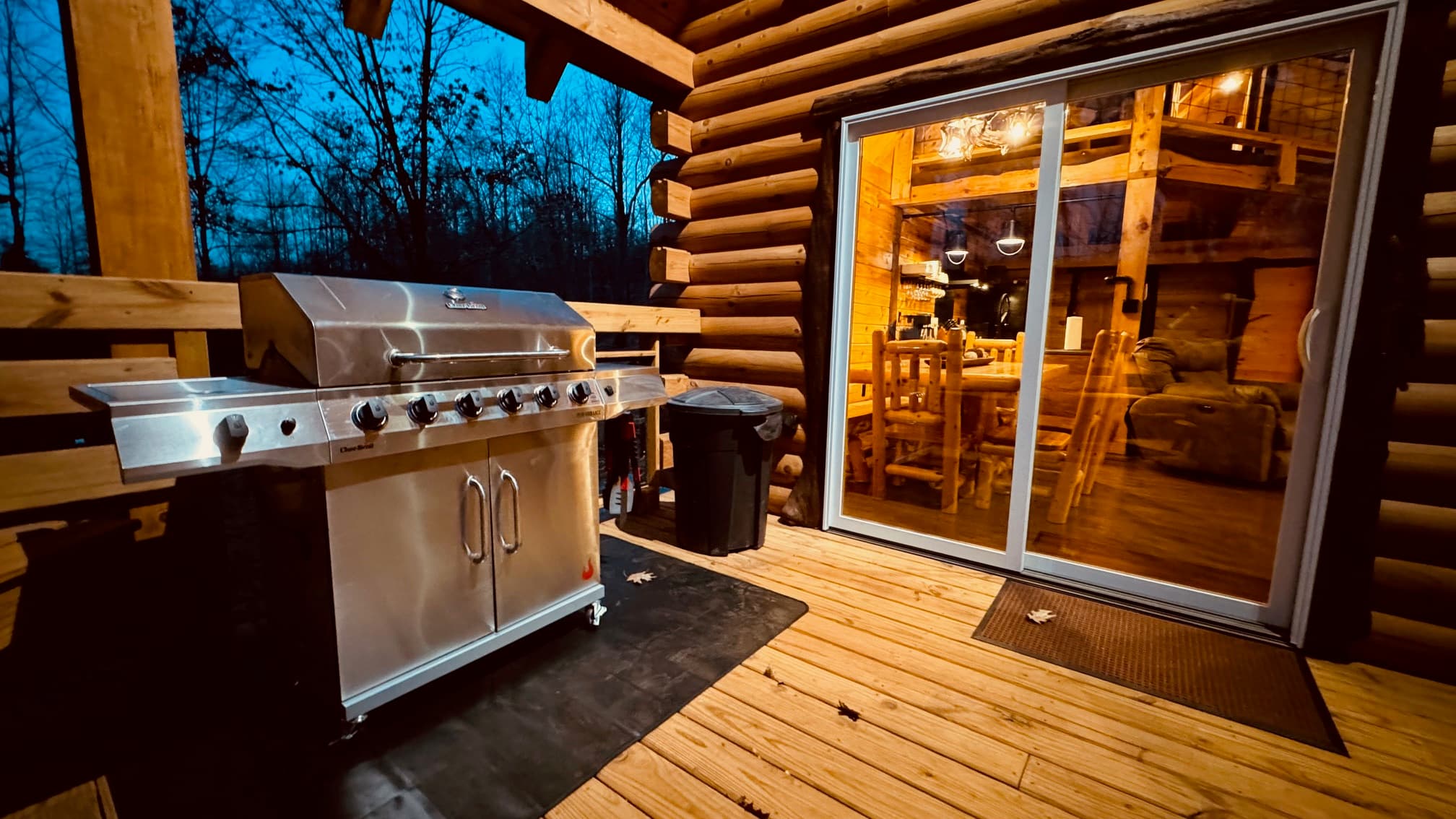 A stainless steel grill sits on a wooden deck outside a log cabin, with a cozy interior illuminated at dusk.
