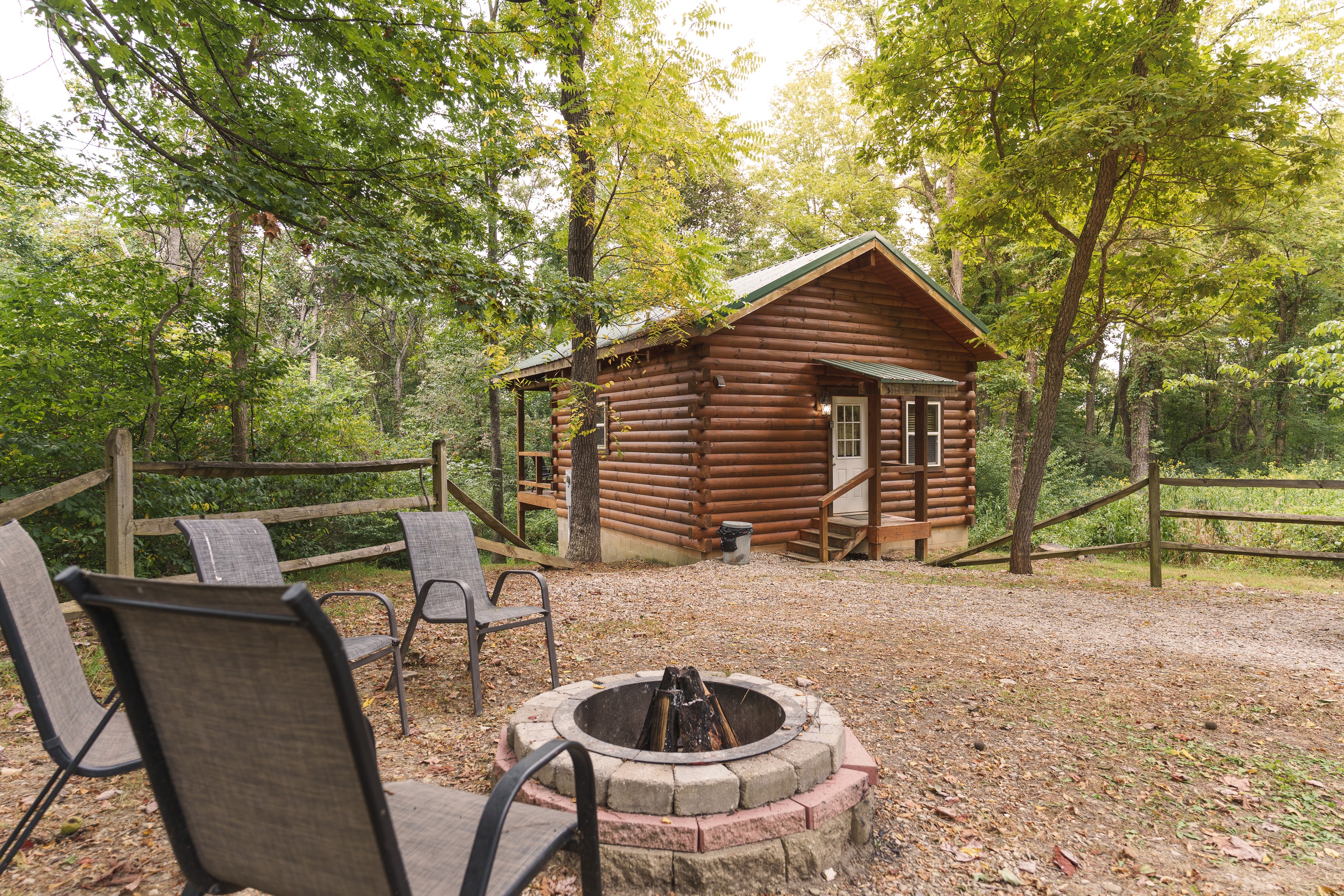 A cozy log cabin surrounded by trees with a fire pit and chairs in the foreground.