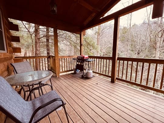 A cozy porch with two chairs, a glass table, and a grill overlooking a wooded area.
