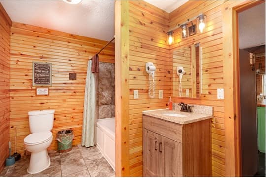 A rustic bathroom featuring wooden walls, a toilet, a bathtub with a shower curtain, and a sink with modern fixtures.