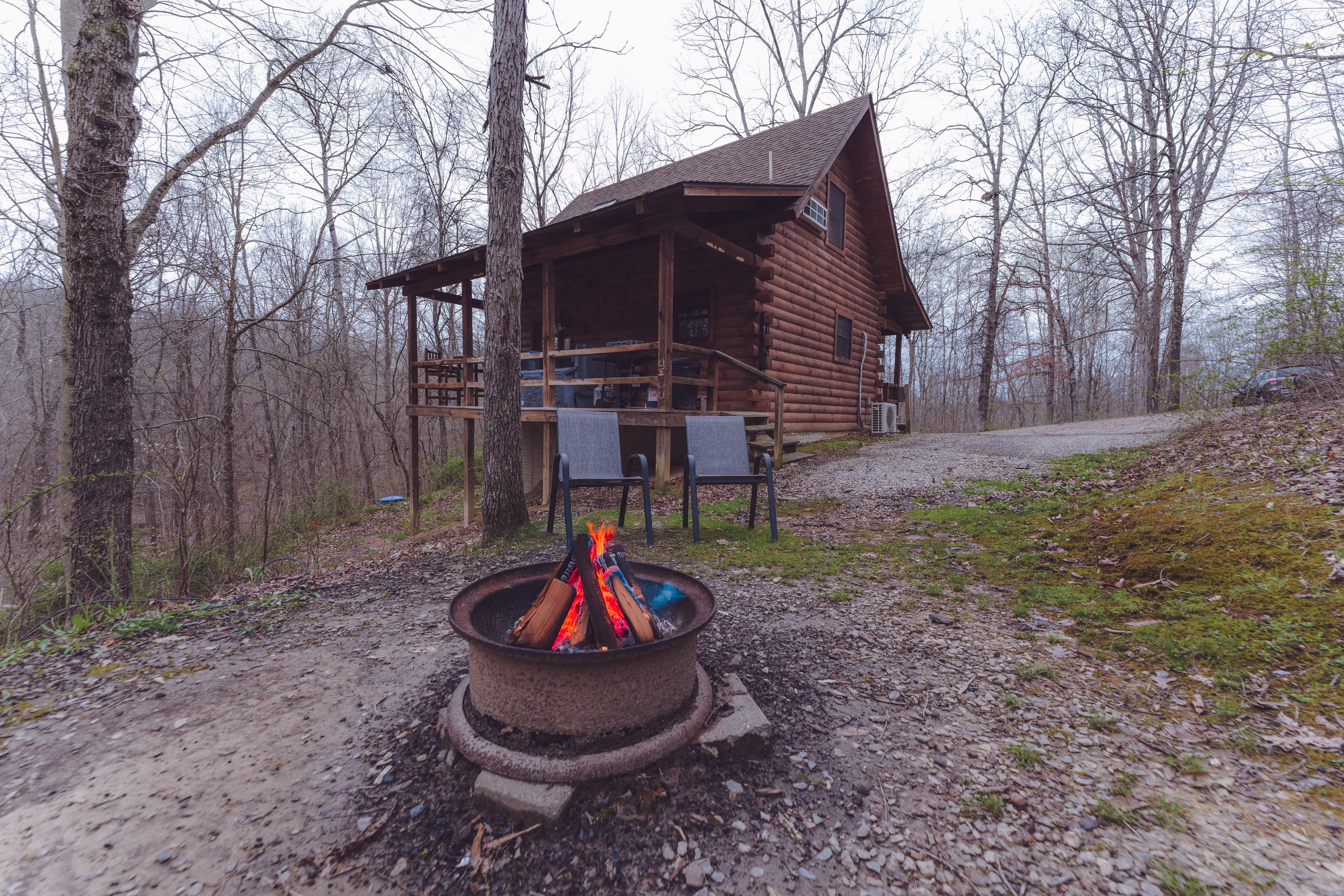 A cozy log cabin surrounded by trees, featuring a campfire pit in the foreground.