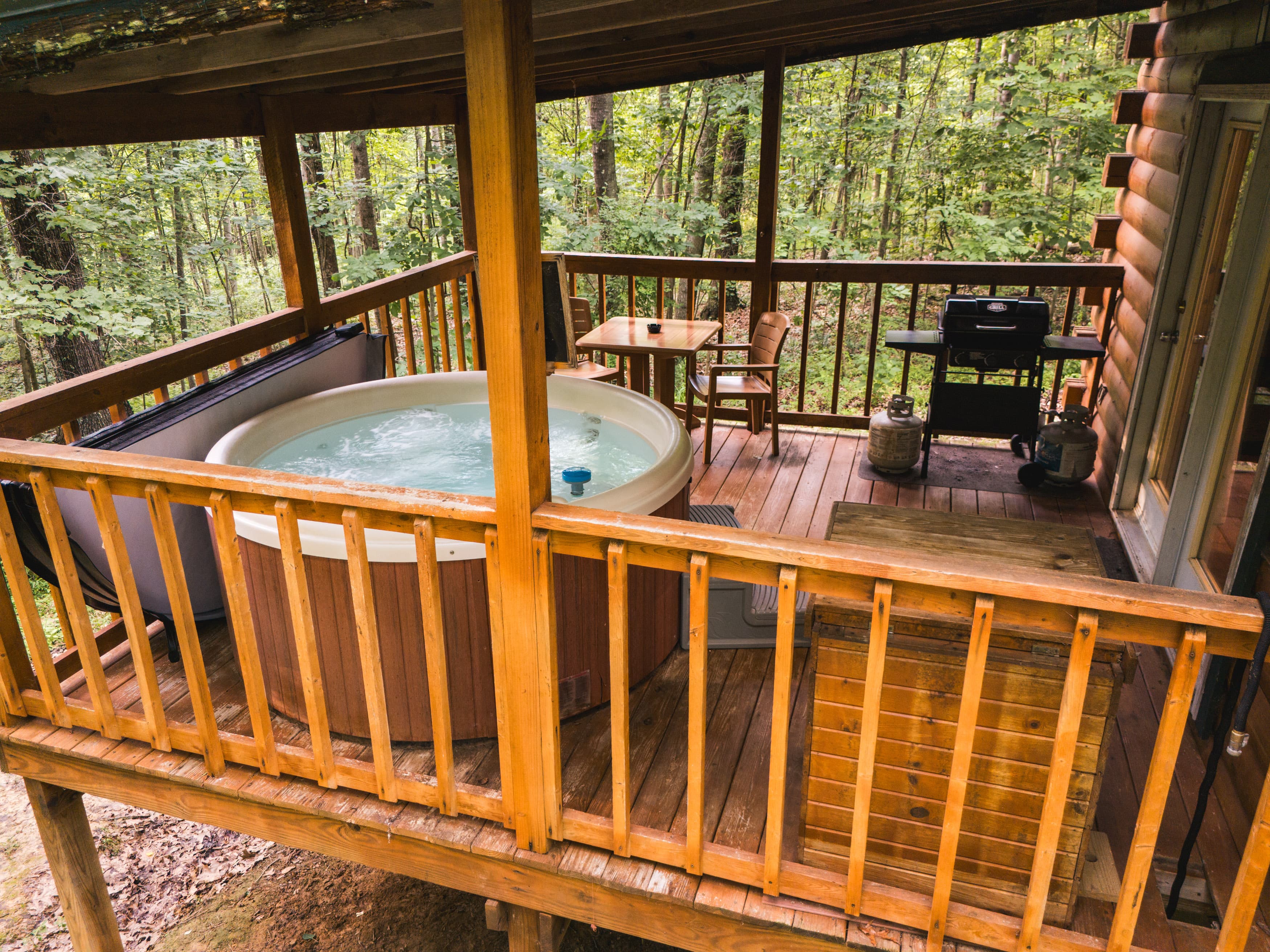 A hot tub and a small outdoor seating area are situated on a wooden deck surrounded by trees.
