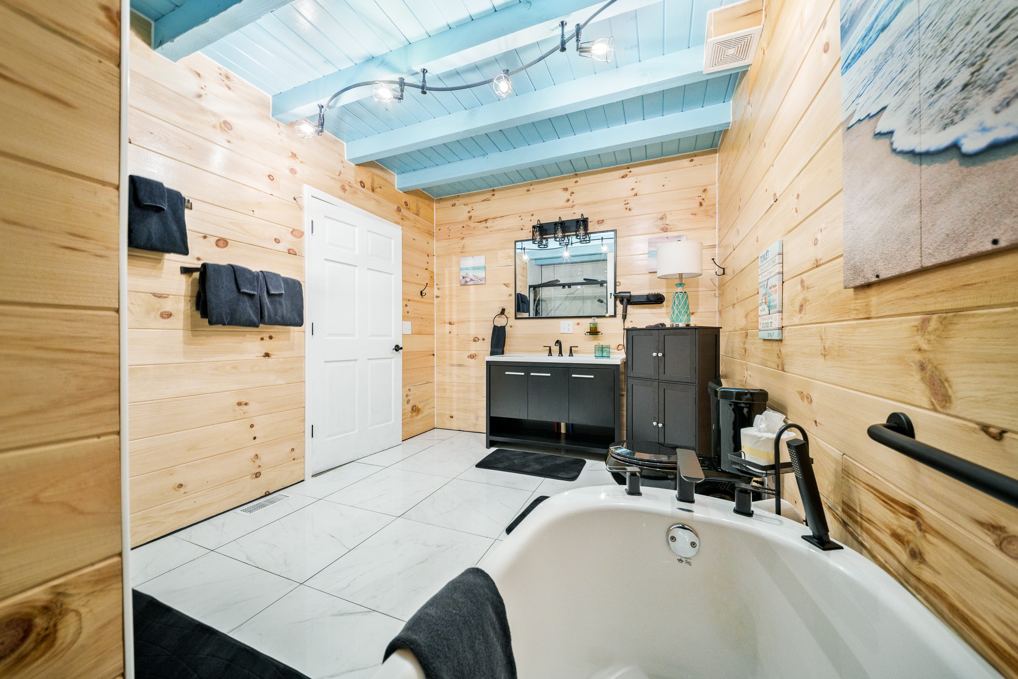 A modern bathroom featuring wooden walls, a freestanding tub, a dark vanity, and blue-accented ceiling.