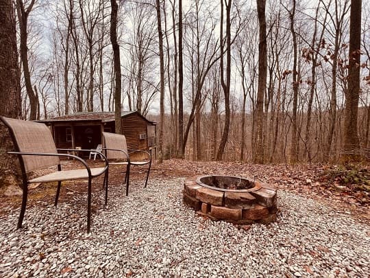 Two chairs beside a stone fire pit in a wooded area with bare trees.