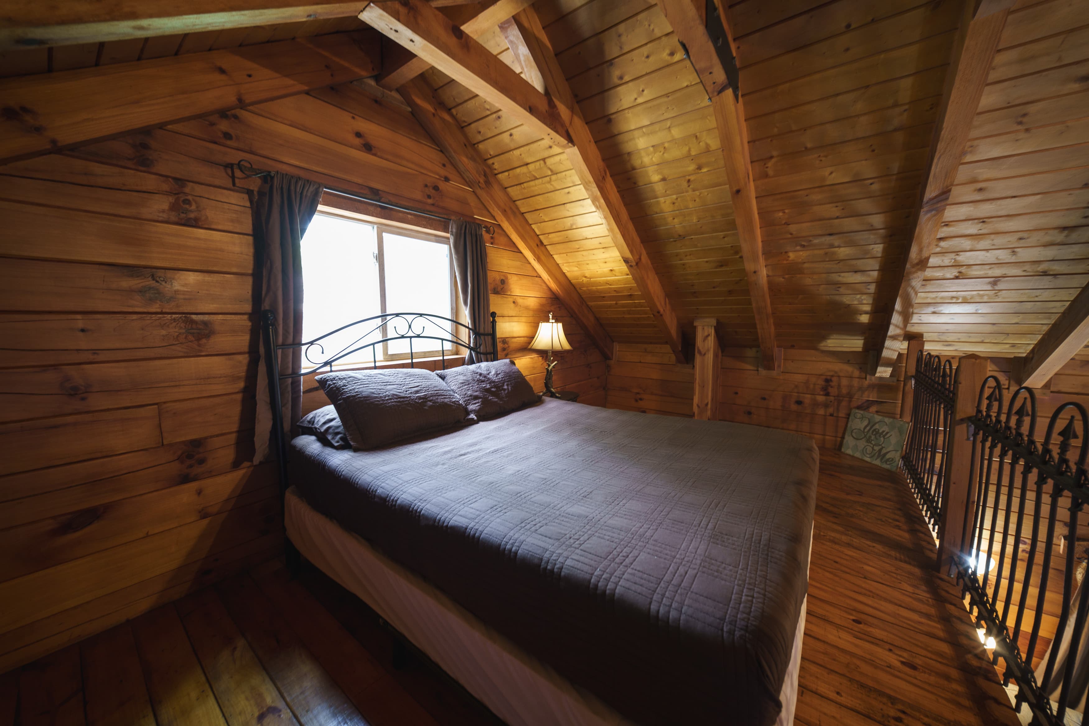 A cozy attic bedroom with a wooden interior, featuring a bed, window, and bedside lamp.