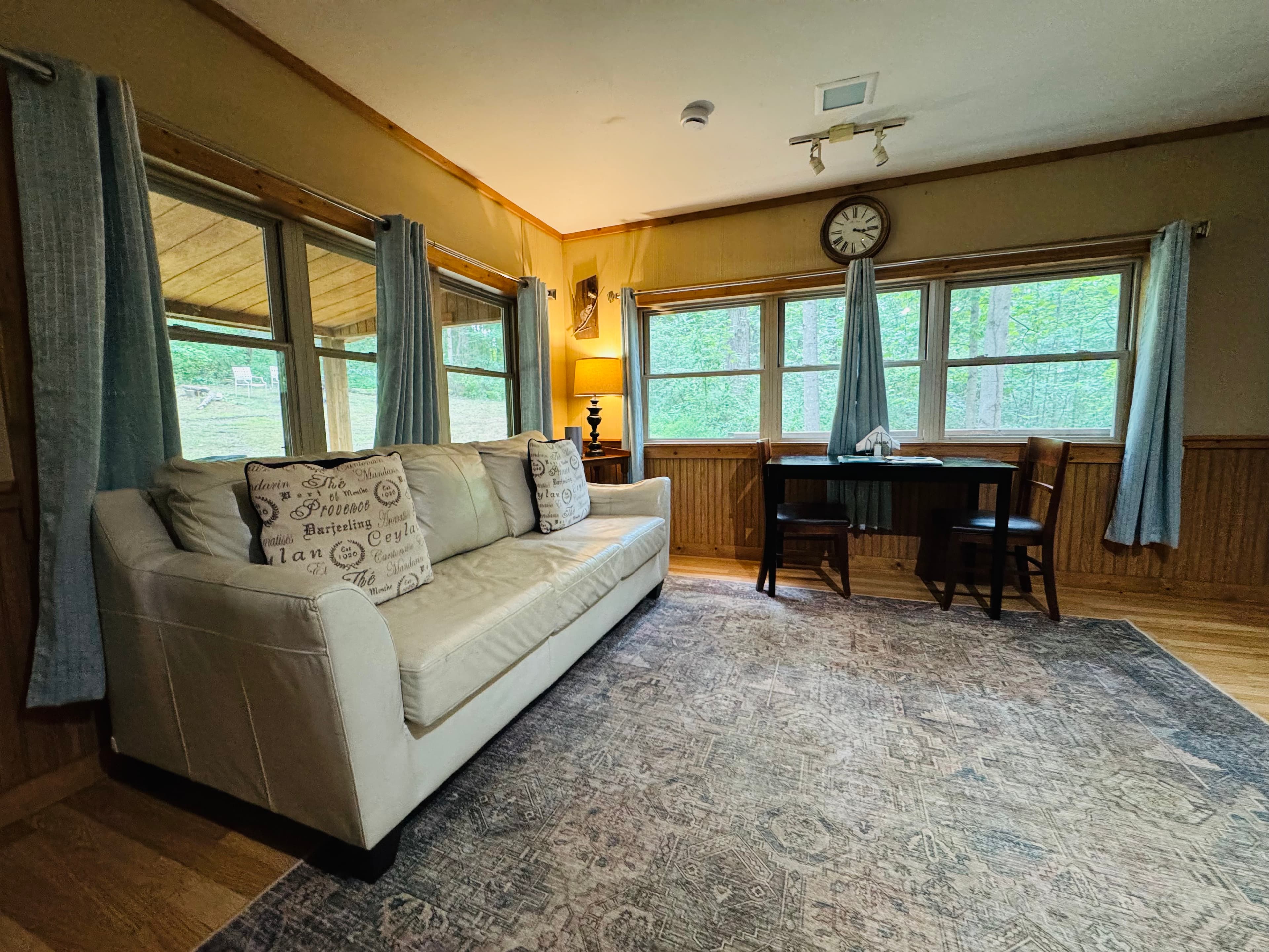 A cozy living space with a white sofa, wooden desk, and large windows overlooking greenery.
