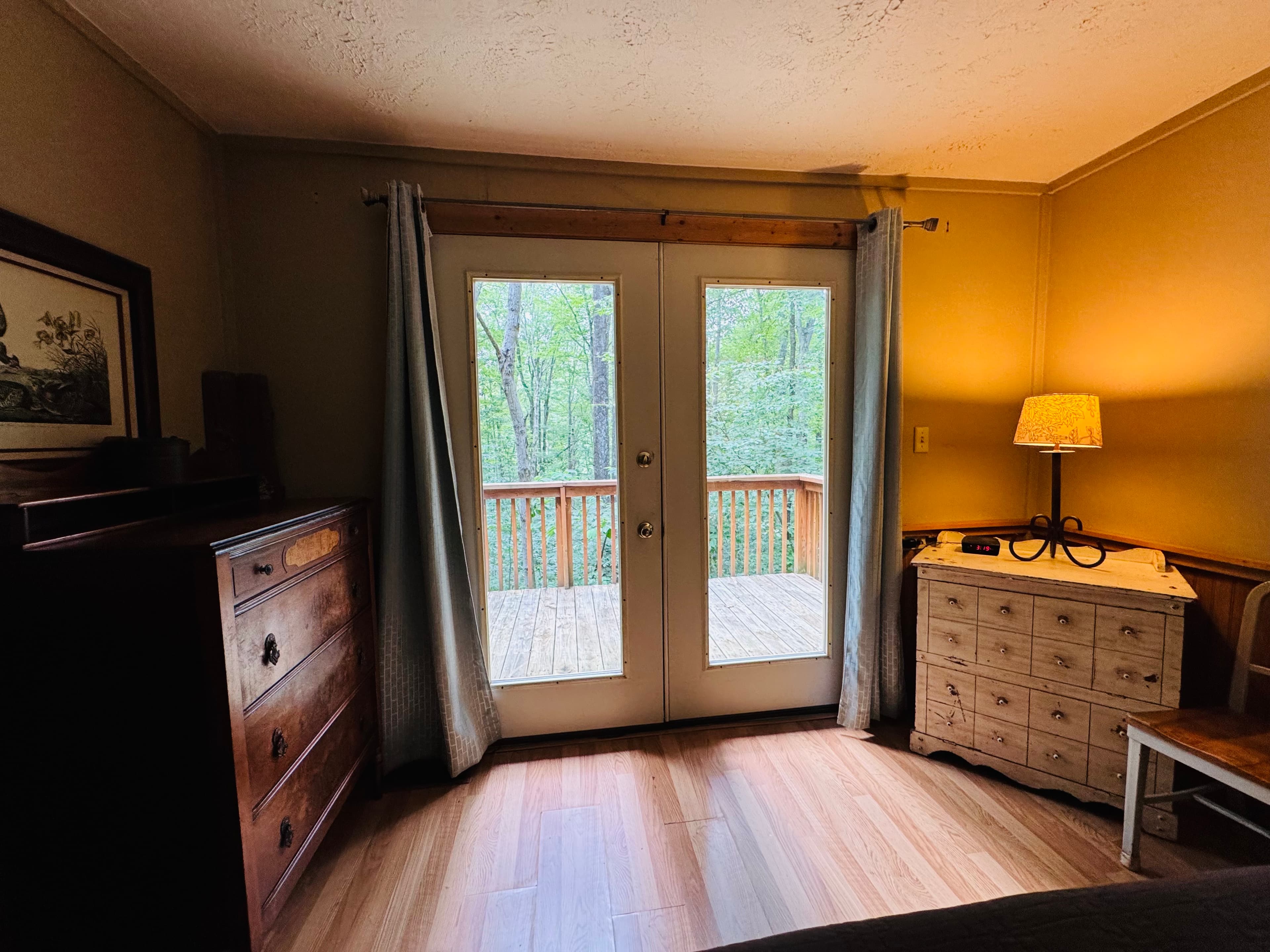 Cozy bedroom with wooden furniture and large doors opening to a deck surrounded by trees.