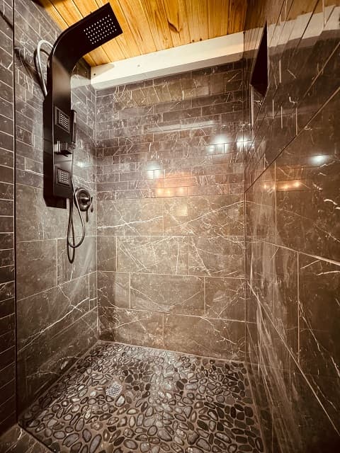 A modern shower with dark marble tiles and a pebble stone floor.
