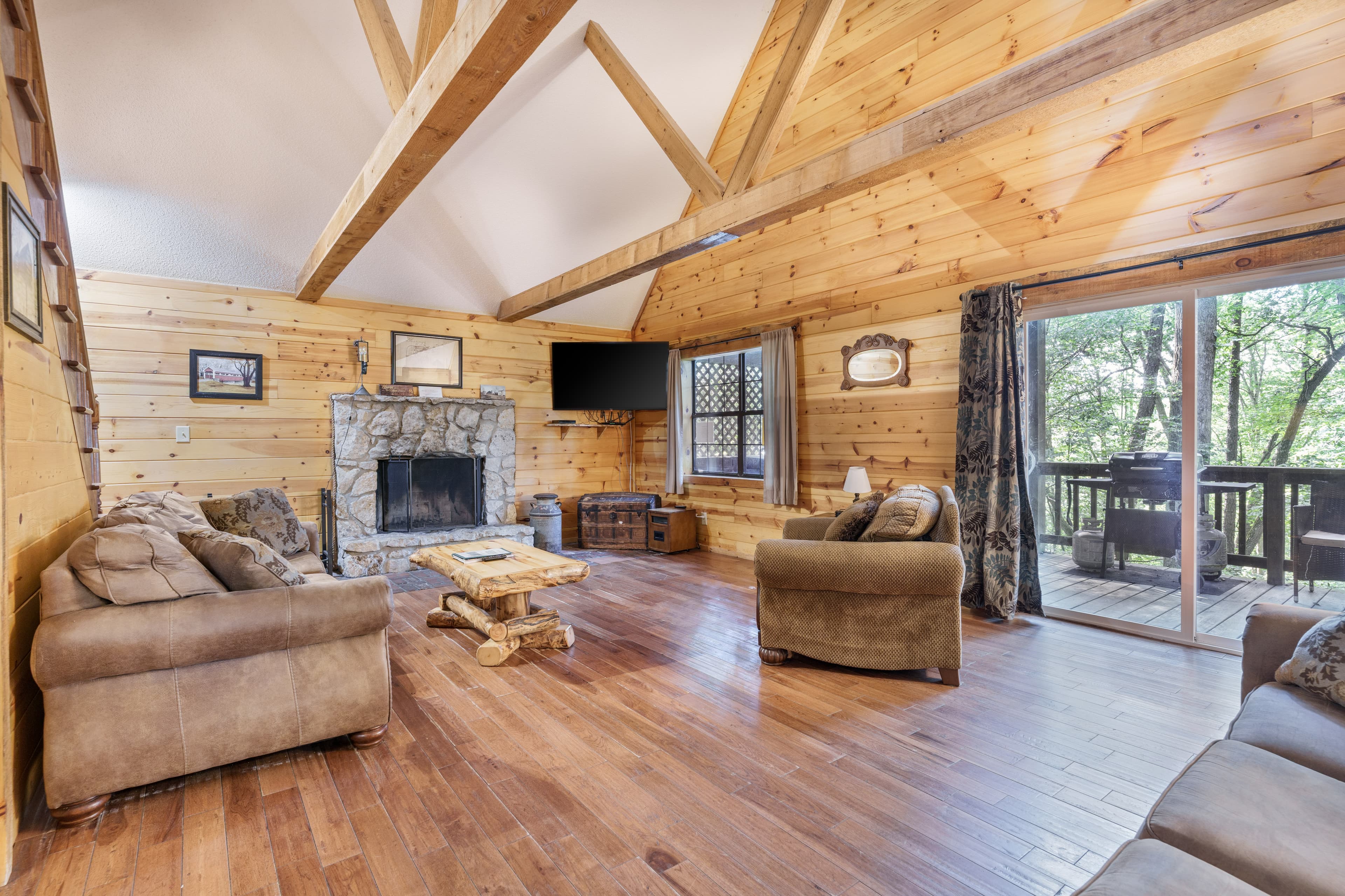 Cozy wooden cabin interior with stone fireplace, plush furniture, and large windows facing a forested view.
