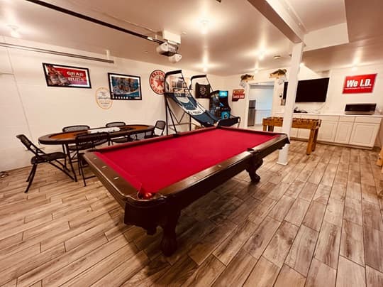 A spacious game room featuring a red pool table, dining area, arcade games, and a mounted TV.