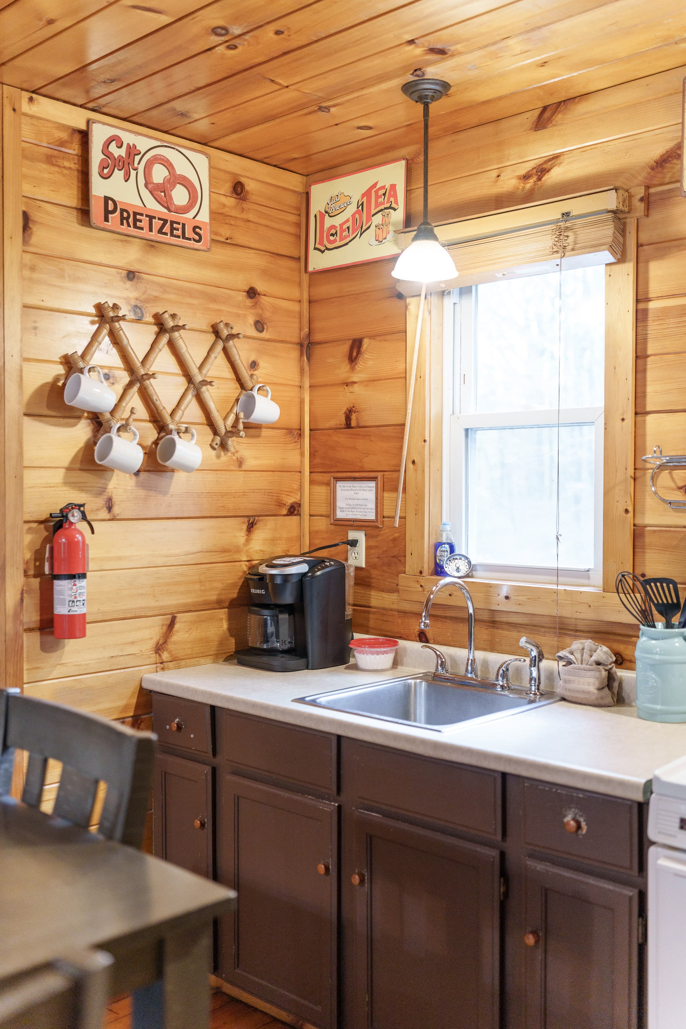 A cozy kitchen corner features wooden walls, a sink, coffee maker, hanging mugs, and vintage signs.