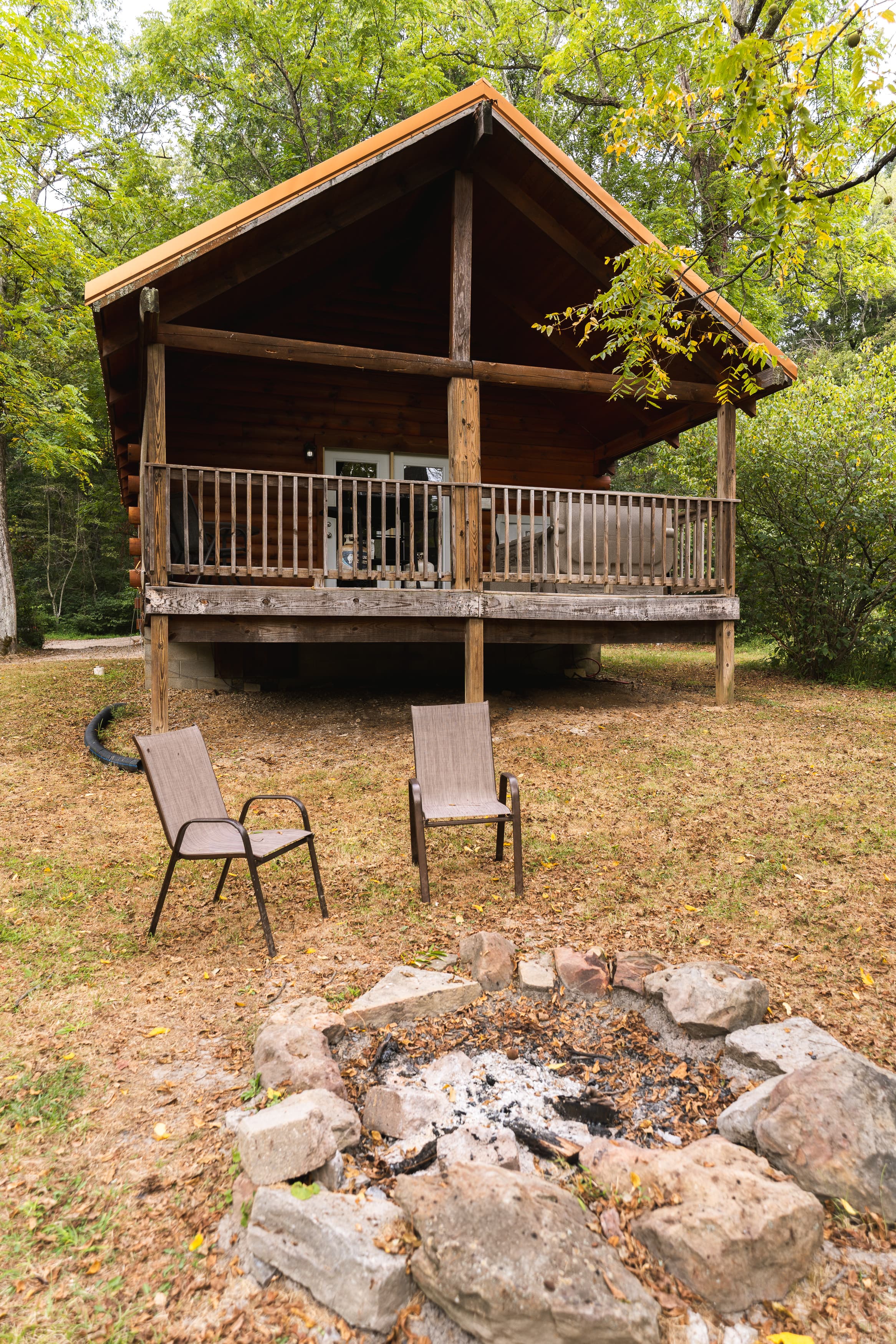 A rustic wooden cabin with a porch, flanked by two chairs and a fire pit in a wooded area.
