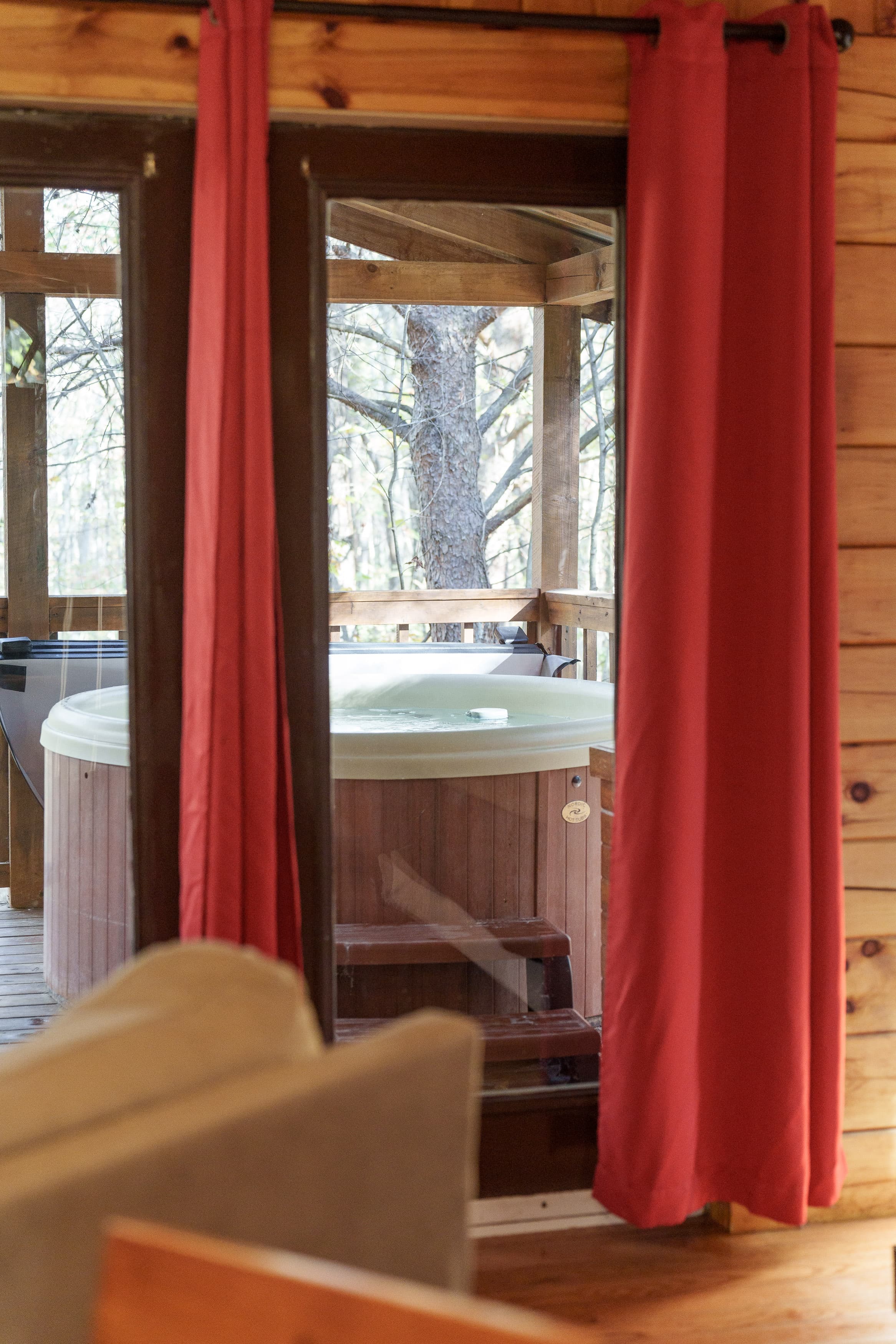 A view through a window with red curtains reveals a hot tub on a wooden deck surrounded by trees.