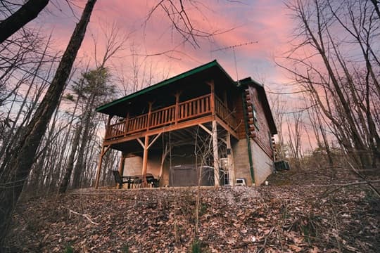 A wooden cabin with a porch sits amidst bare trees under a colorful sunset sky.