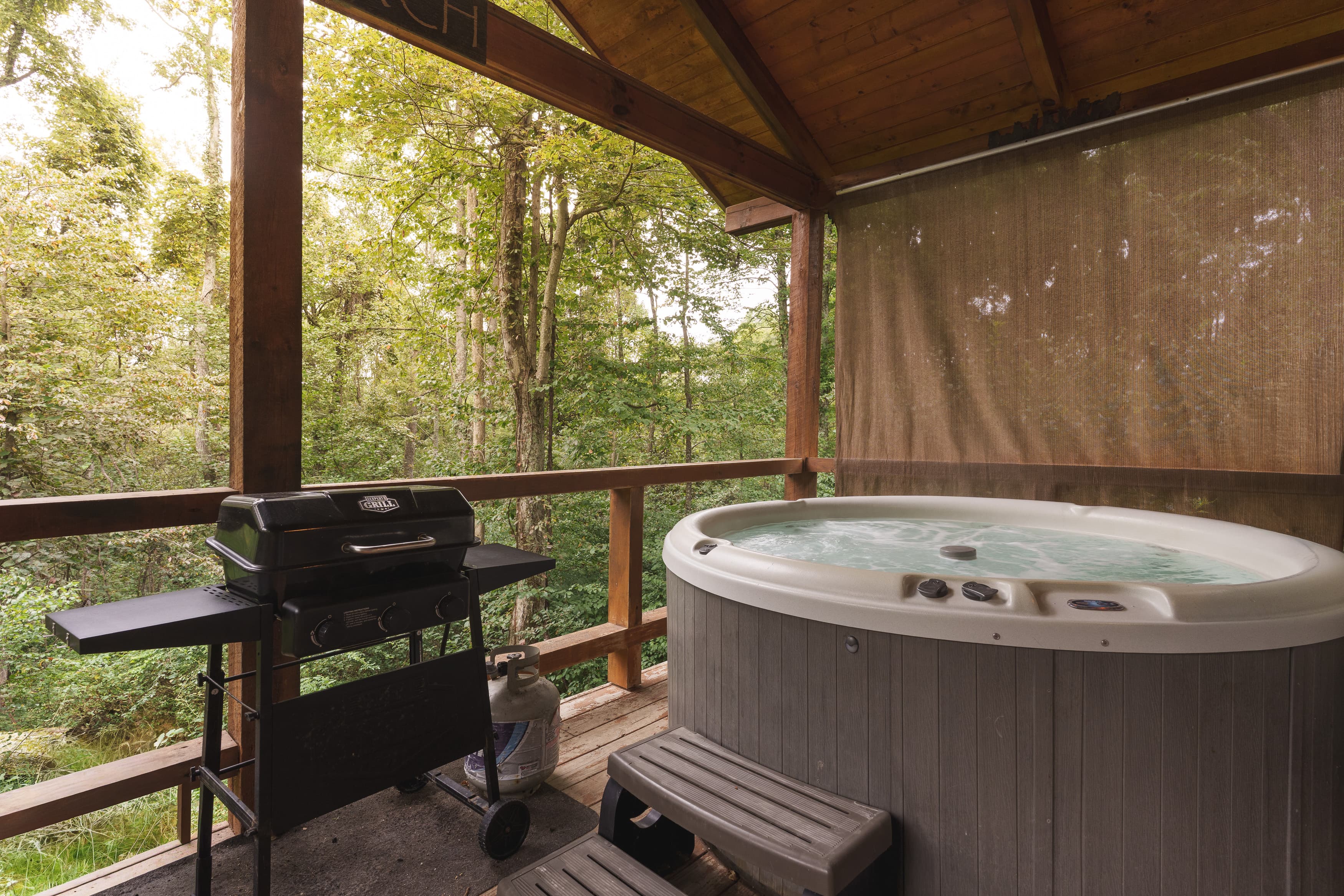 A hot tub and grill are situated on a wooden deck surrounded by trees.