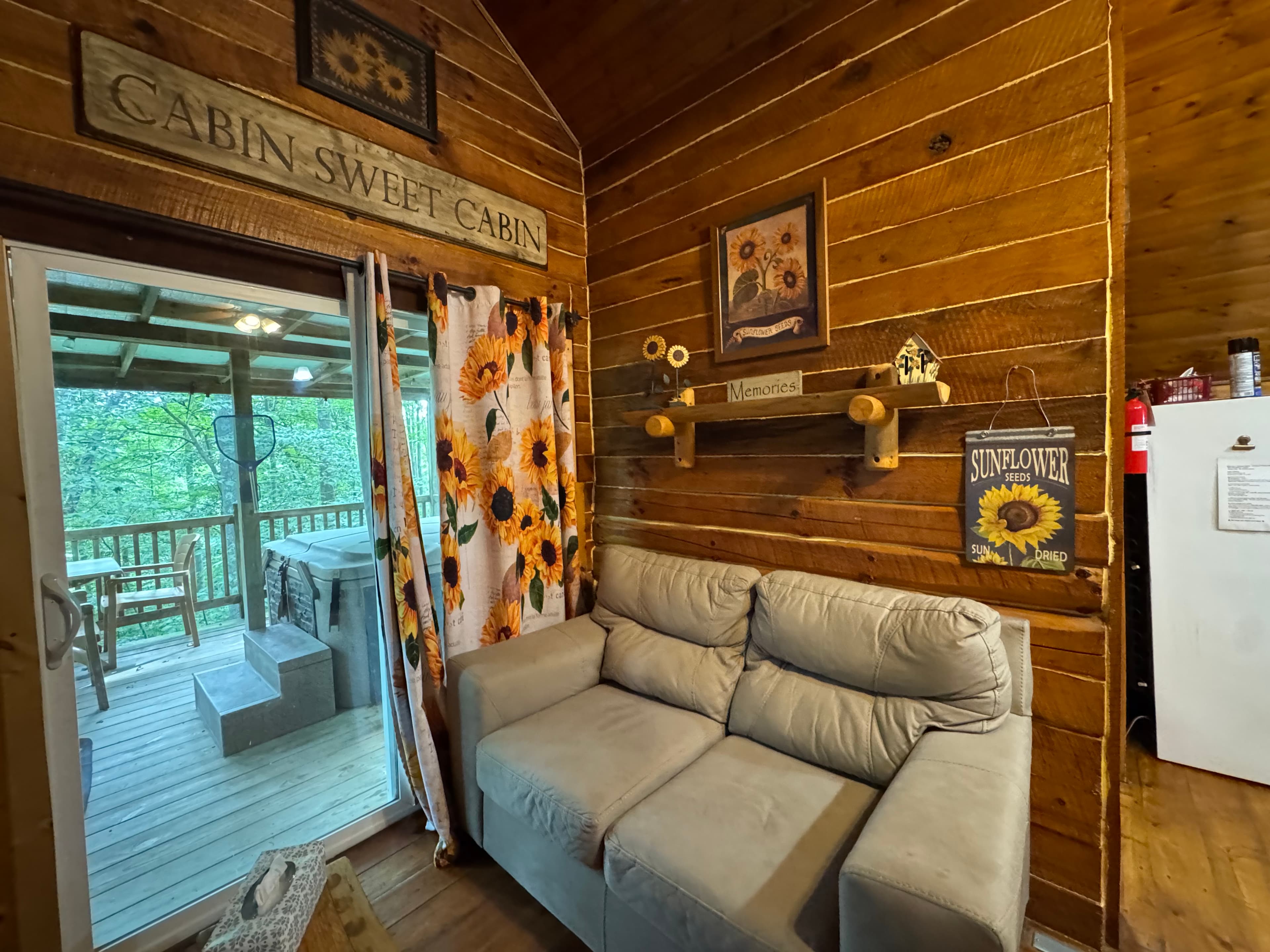 Cozy cabin interior featuring a gray sofa, sunflower-themed decor, and a view of a deck through large glass doors.