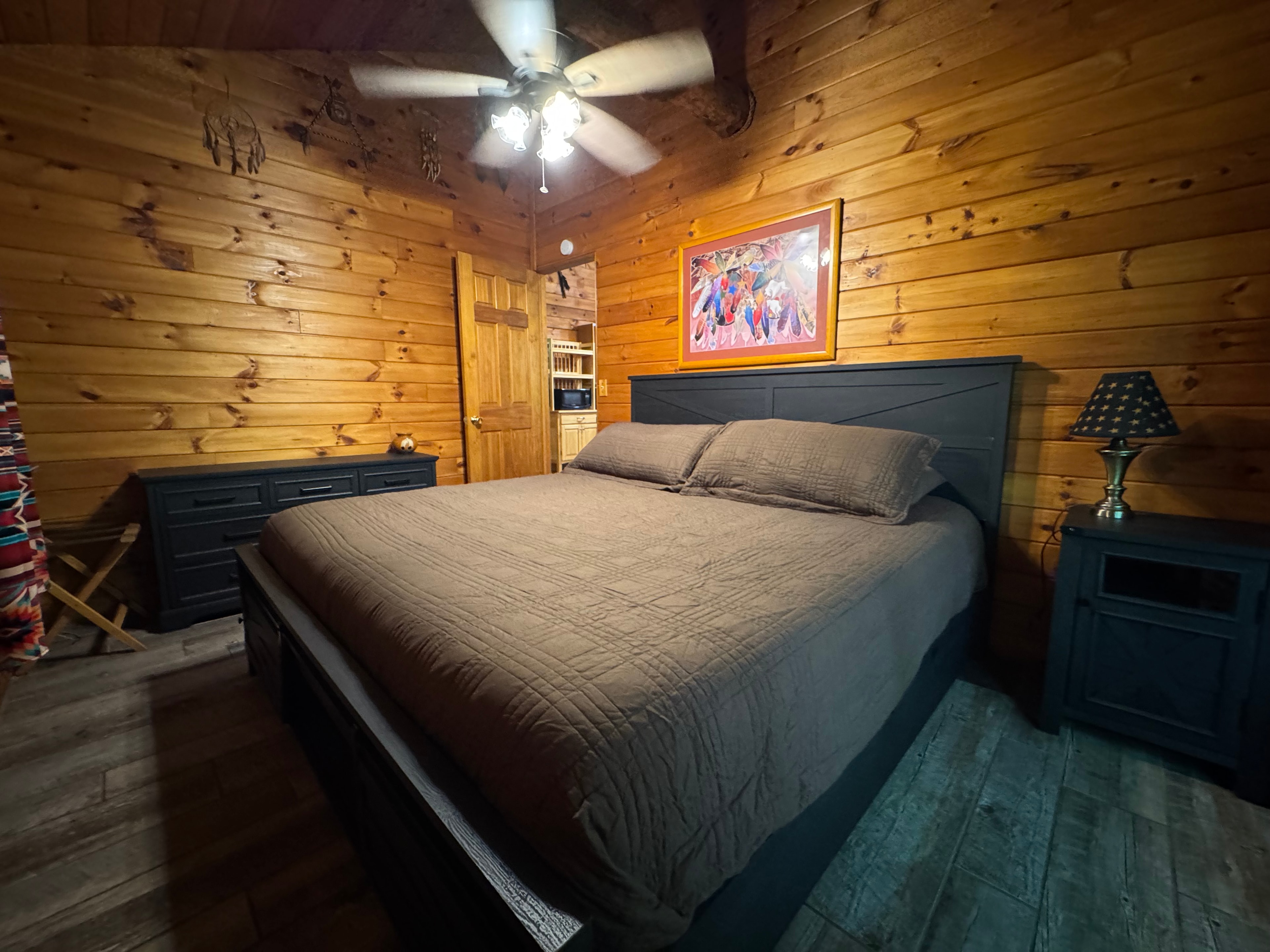 A cozy bedroom featuring a dark wooden bed, dresser, and ceiling fan against a backdrop of wooden walls.