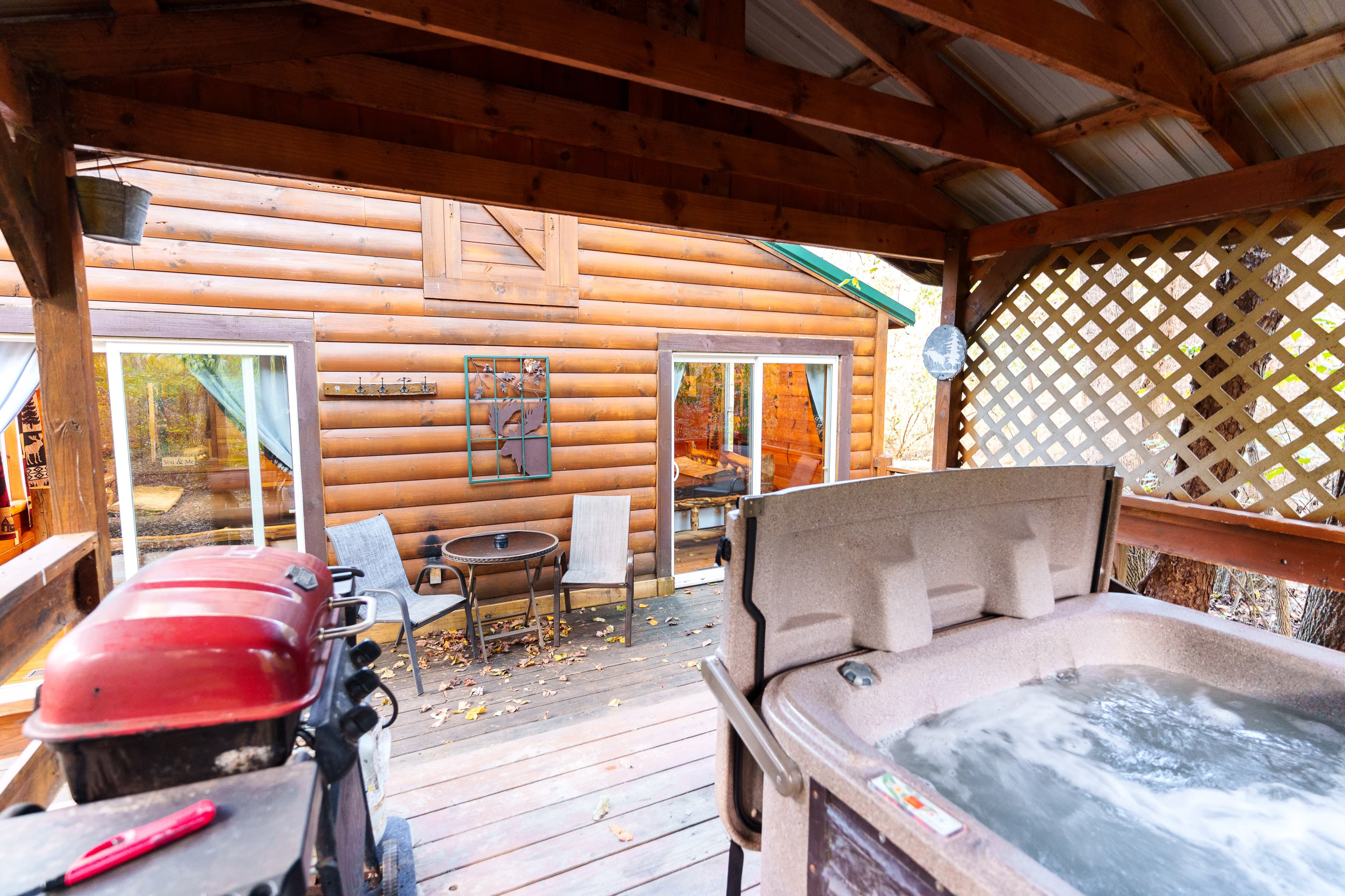 A wooden deck featuring a hot tub and a grill, with a cabin in the background and chairs arranged by a table.
