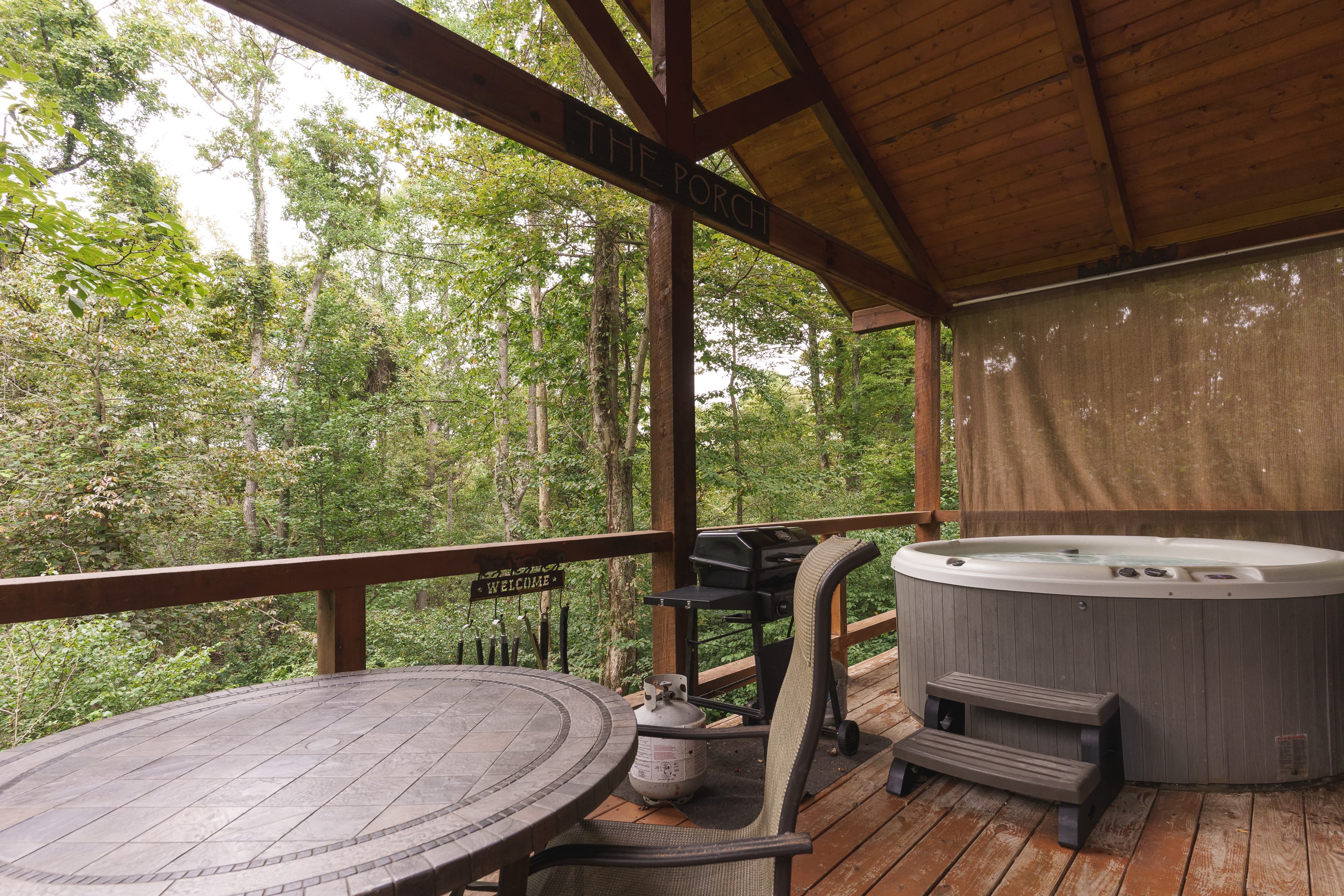 A cozy outdoor porch with a hot tub, dining table, and grill, surrounded by lush greenery.