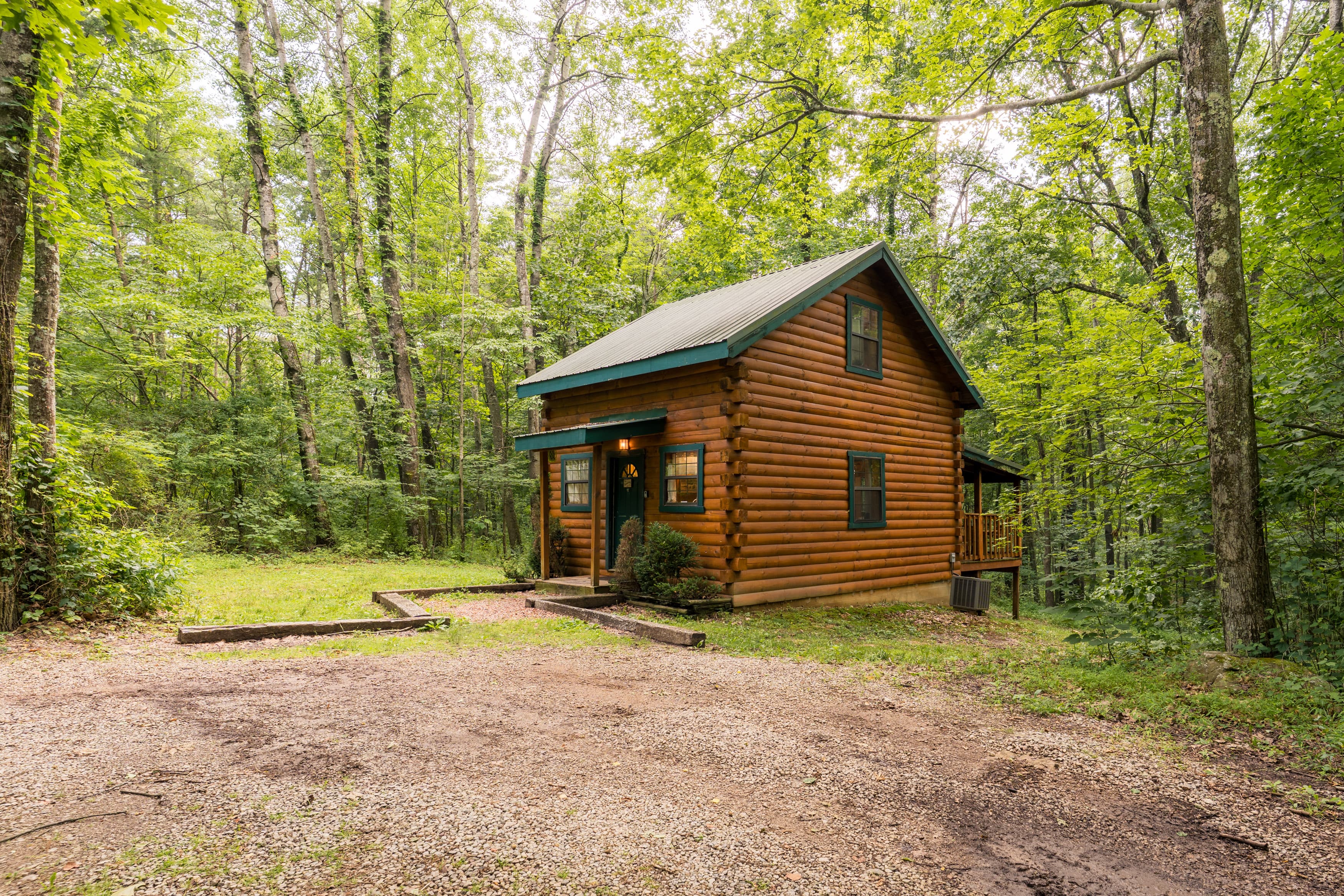A cozy log cabin surrounded by lush green trees and a gravel driveway.