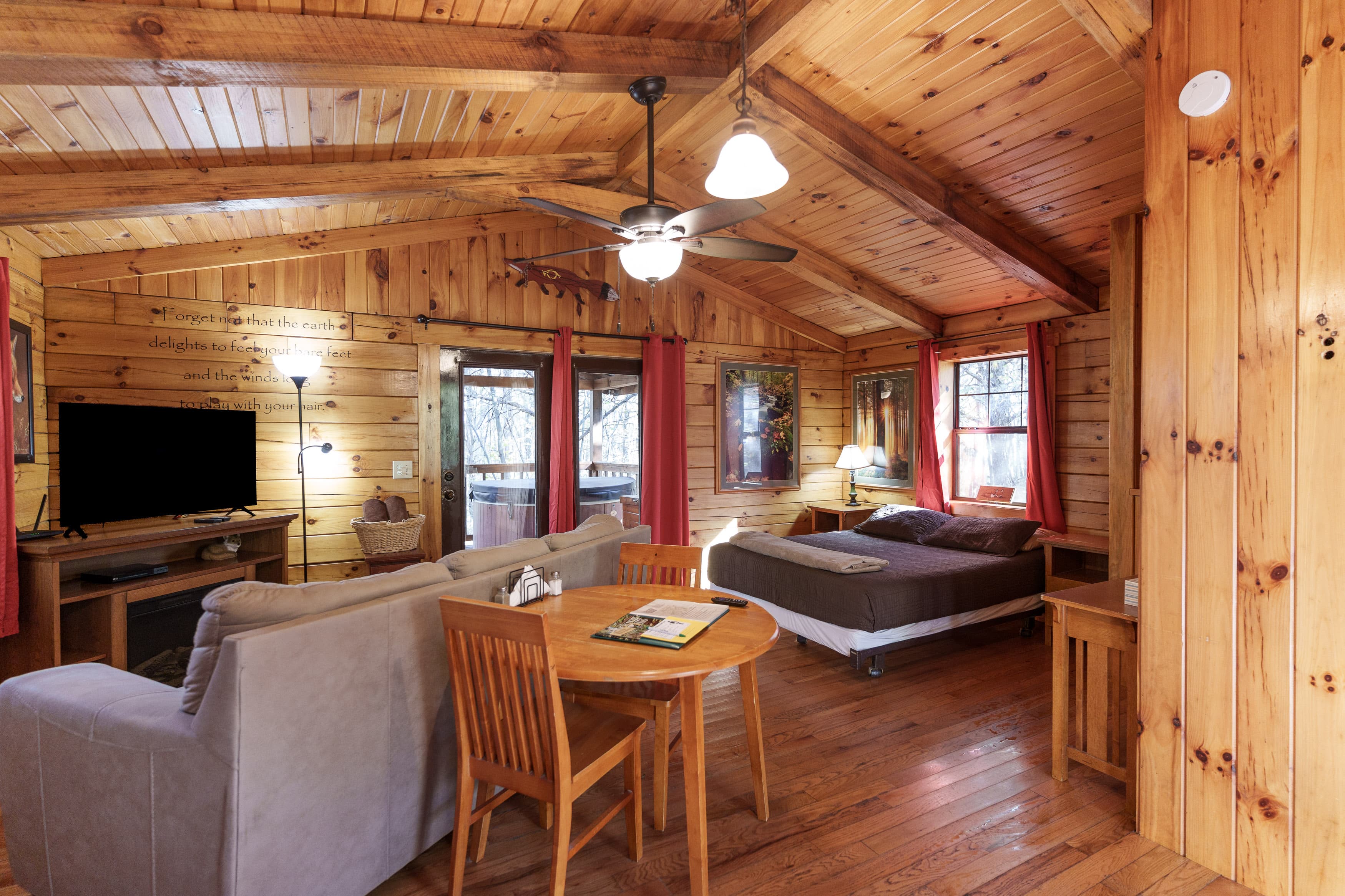 Cozy wooden cabin interior featuring a living area, dining table, and a bedroom with large windows.